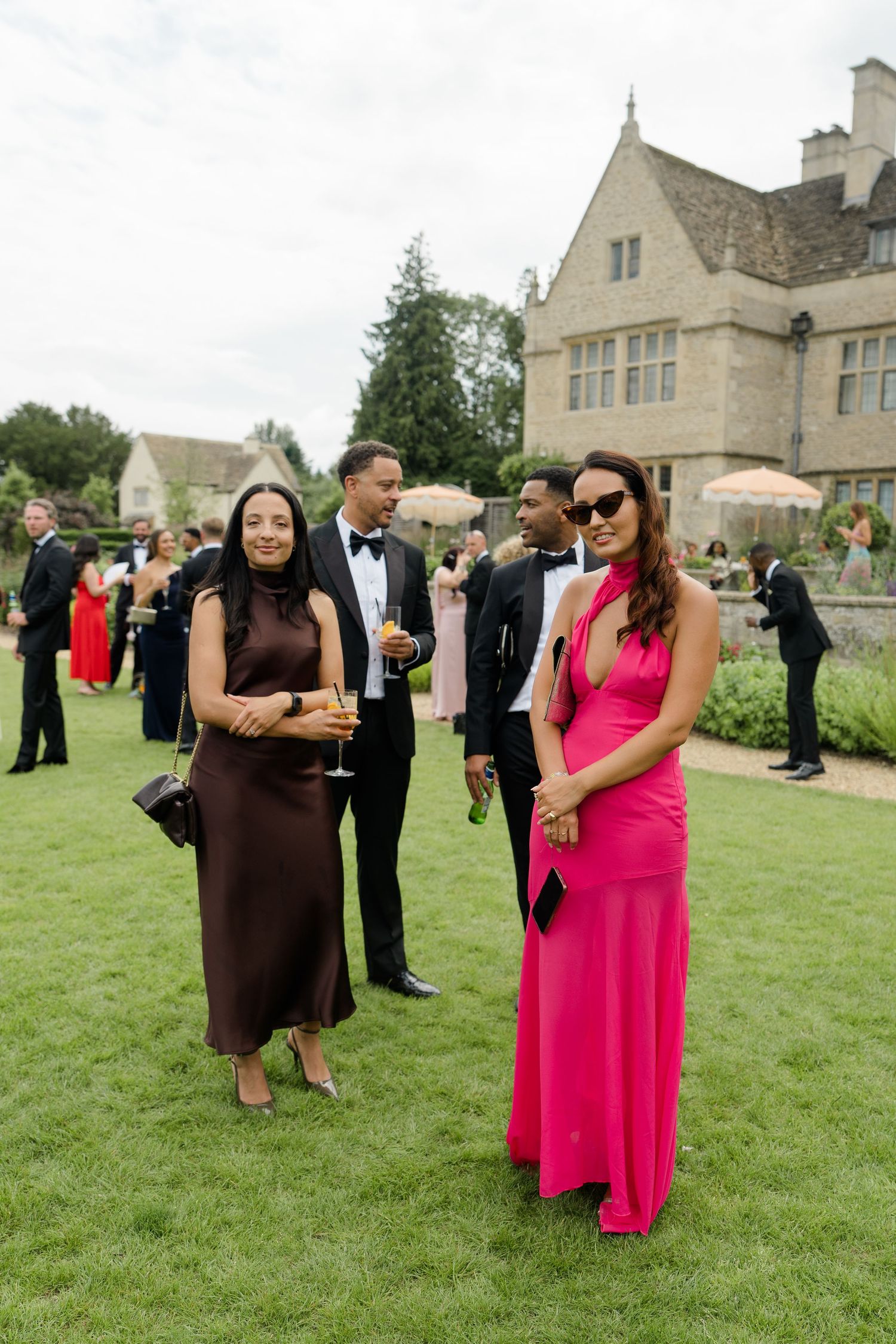 Wedding guests posing on lawn, woman in brown silk dress and woman in hot pink gown at elegant manor house reception.