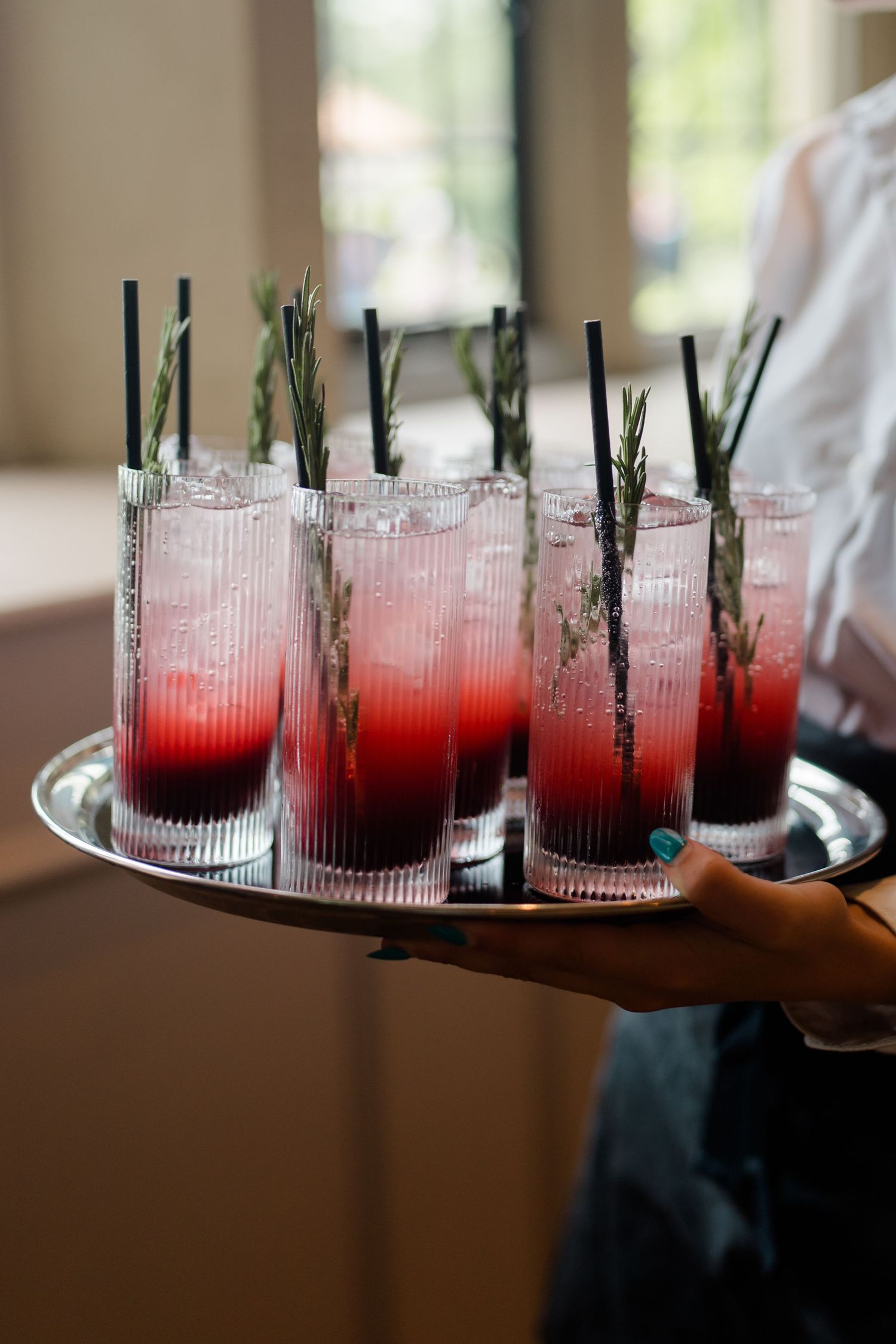 A server carries a tray of red gradient cocktails with rosemary garnishes and black straws at an elegant event.