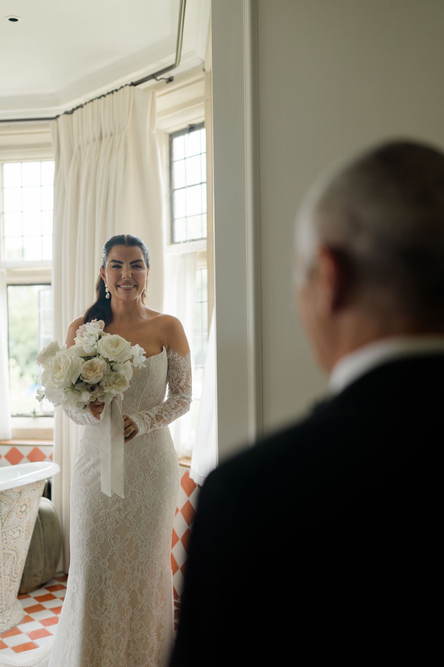 A radiant bride in a lace off-shoulder gown holds white bouquet during an emotional first look moment.