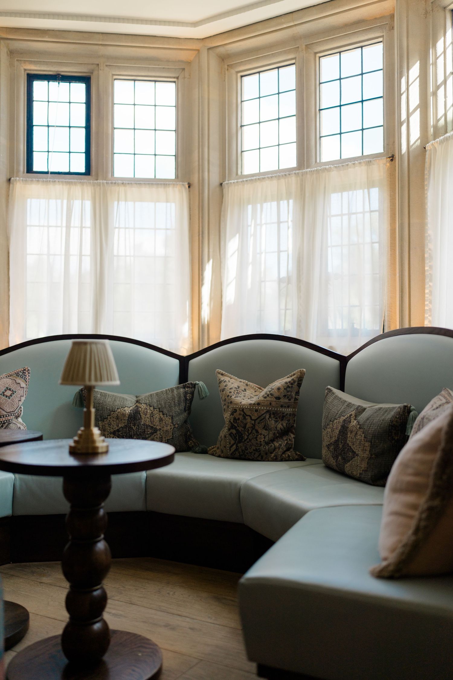 Elegant curved sage green sofa with decorative pillows in a bay window sitting room with sheer curtains.