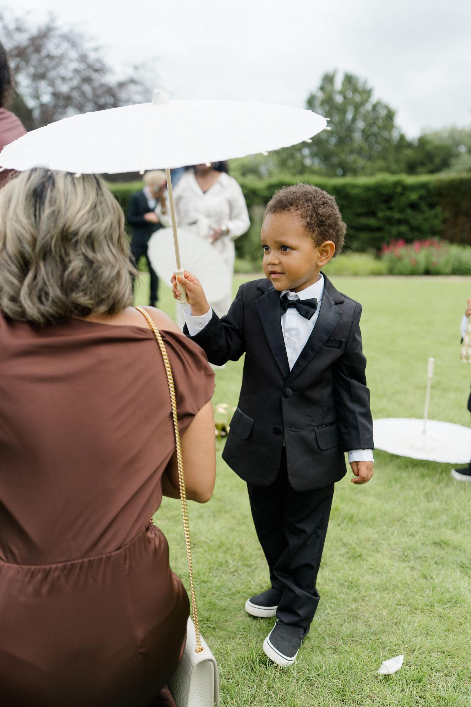 Adorable young ring bearer in black tuxedo holding white parasol at outdoor garden wedding.