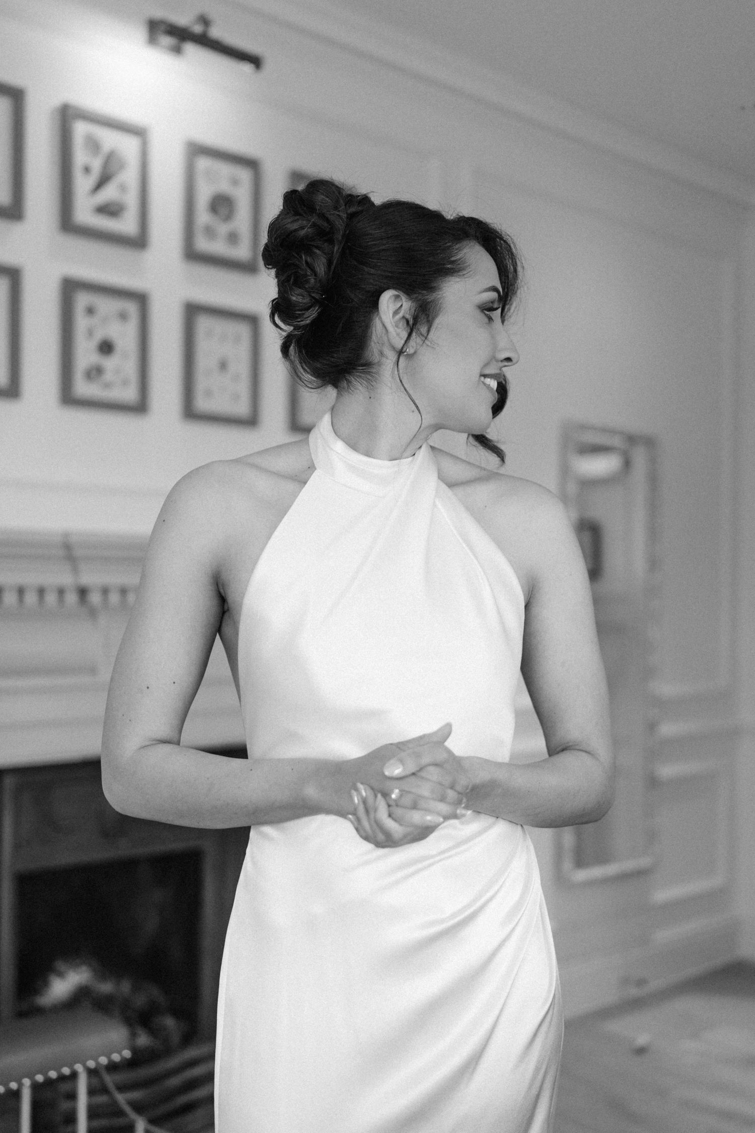 Black and white portrait of bride in halter neck wedding gown with upswept hair by a fireplace.
