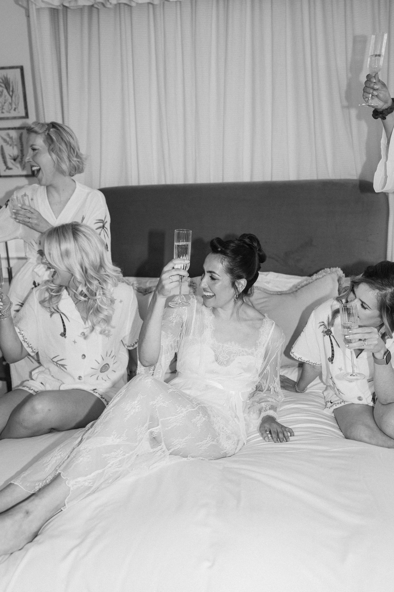 Bride in wedding dress raises champagne glass with bridesmaids in pajamas on bed during bridal prep.