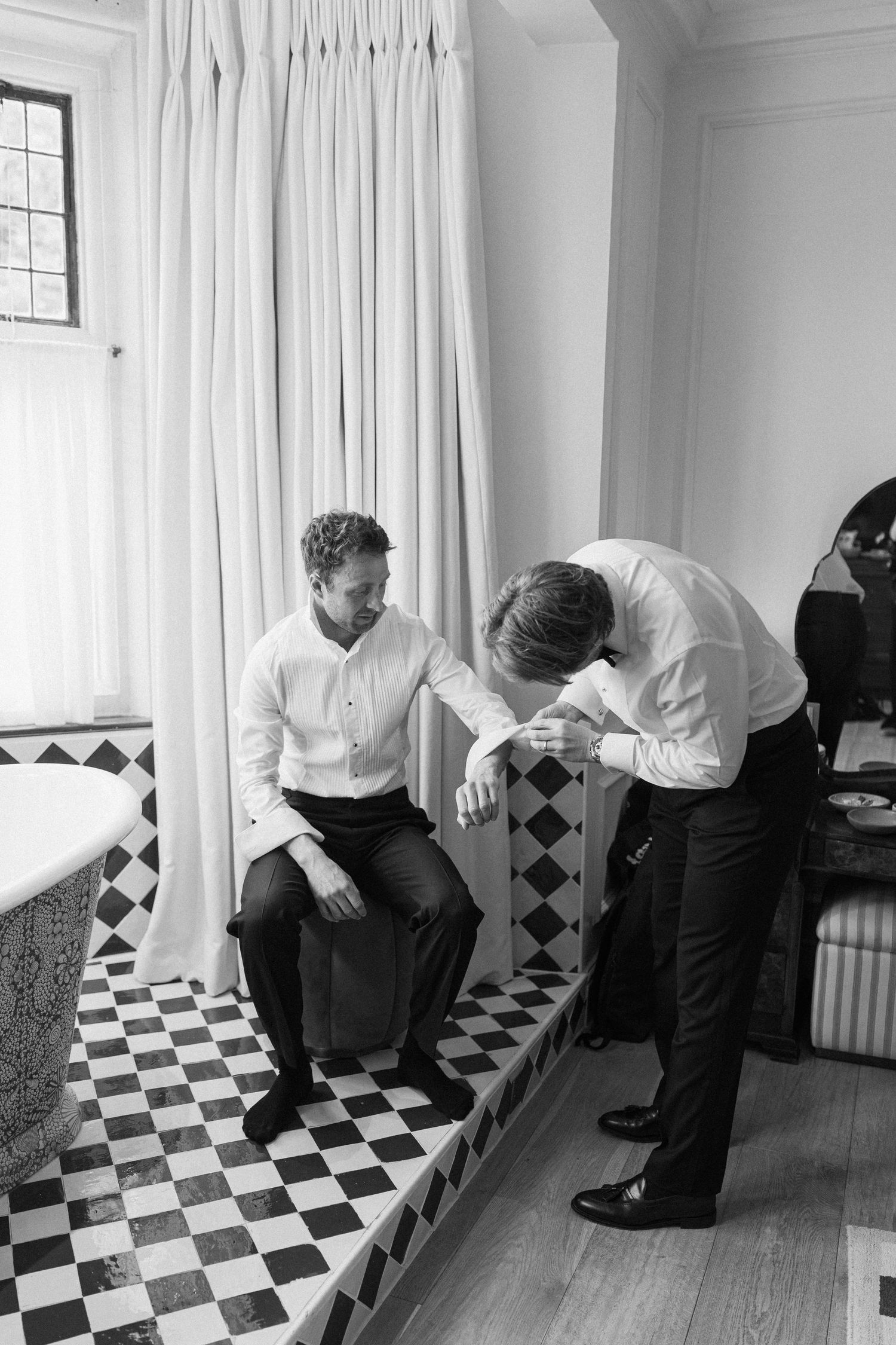 Groom sits on bathroom ledge while groomsman helps adjust cufflinks in black and white wedding photo.