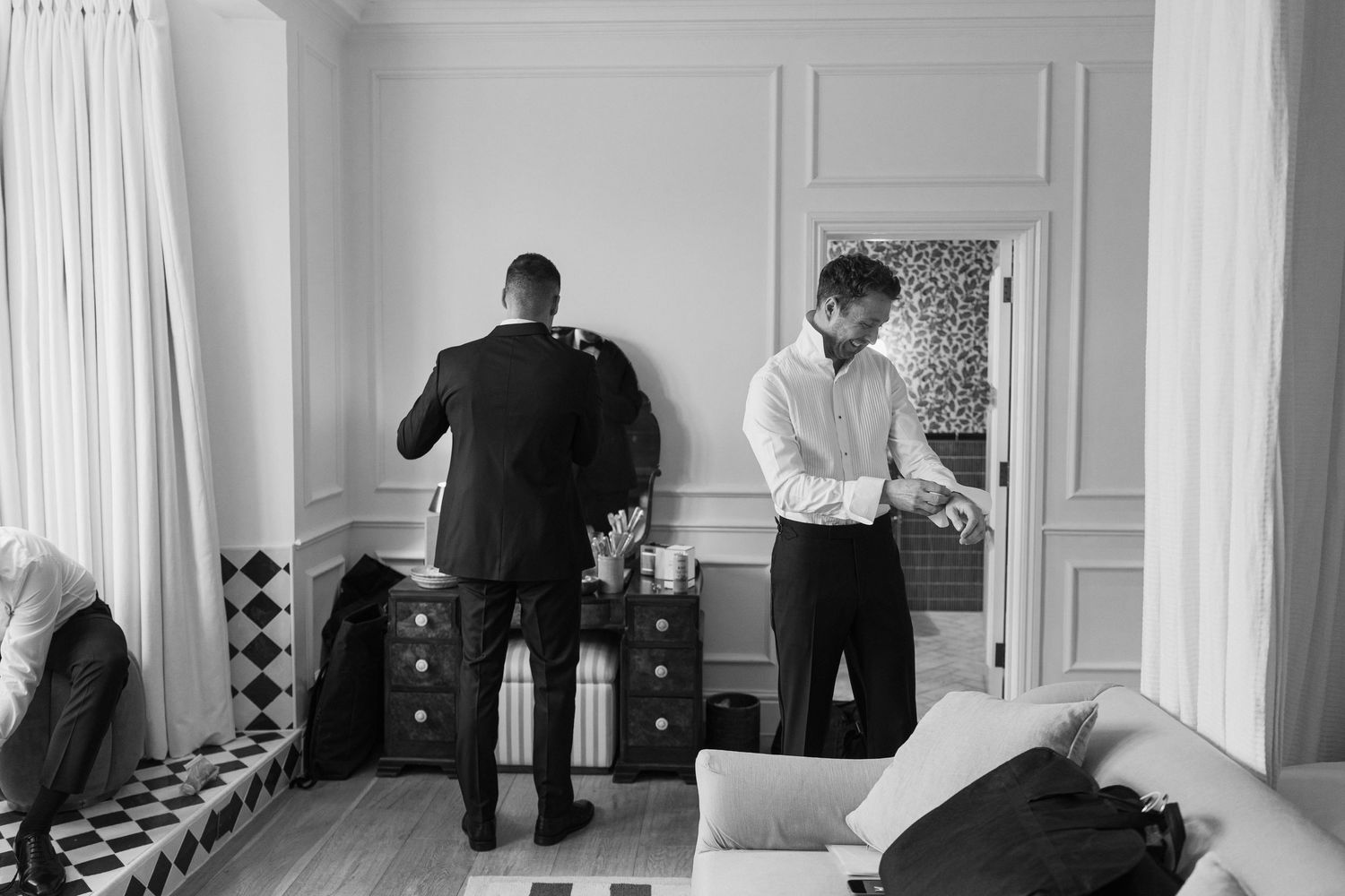 Black and white photo of two groomsmen in tuxedos helping each other get dressed in an elegant hotel room.