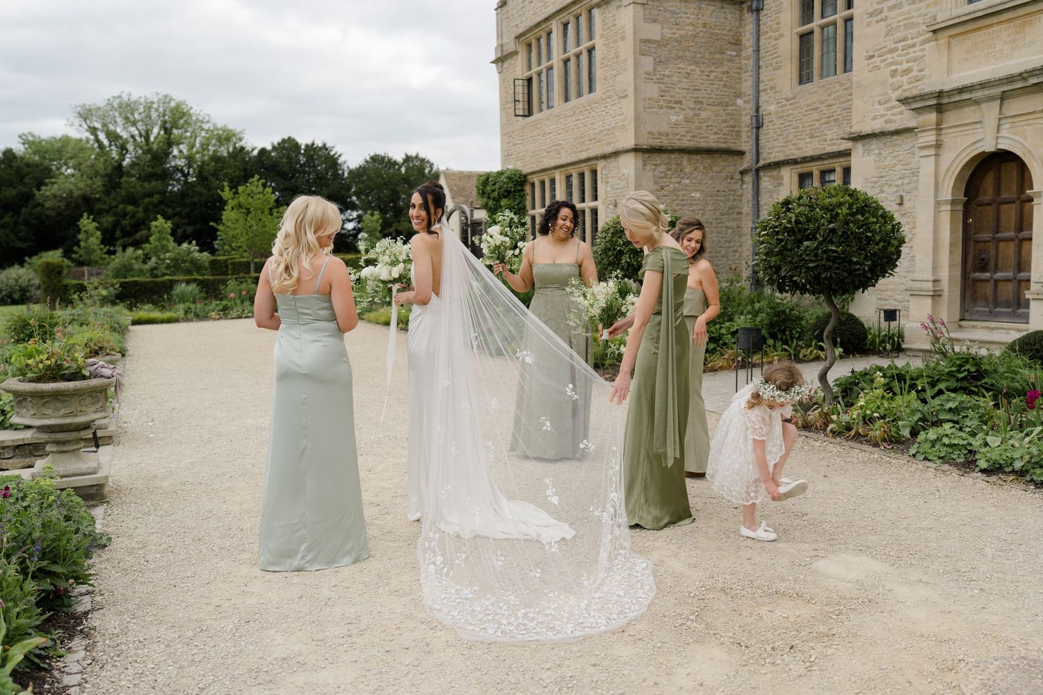 Bride and bridesmaids admiring lace wedding veil in garden of a historic stone manor house.