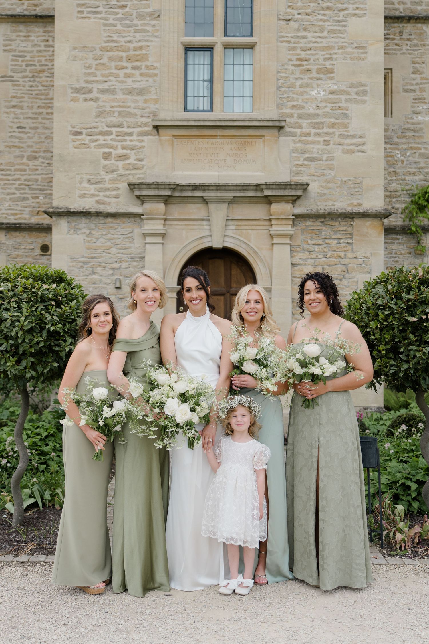 Bride in white halter gown poses with four bridesmaids in sage green dresses and flower girl before stone manor.