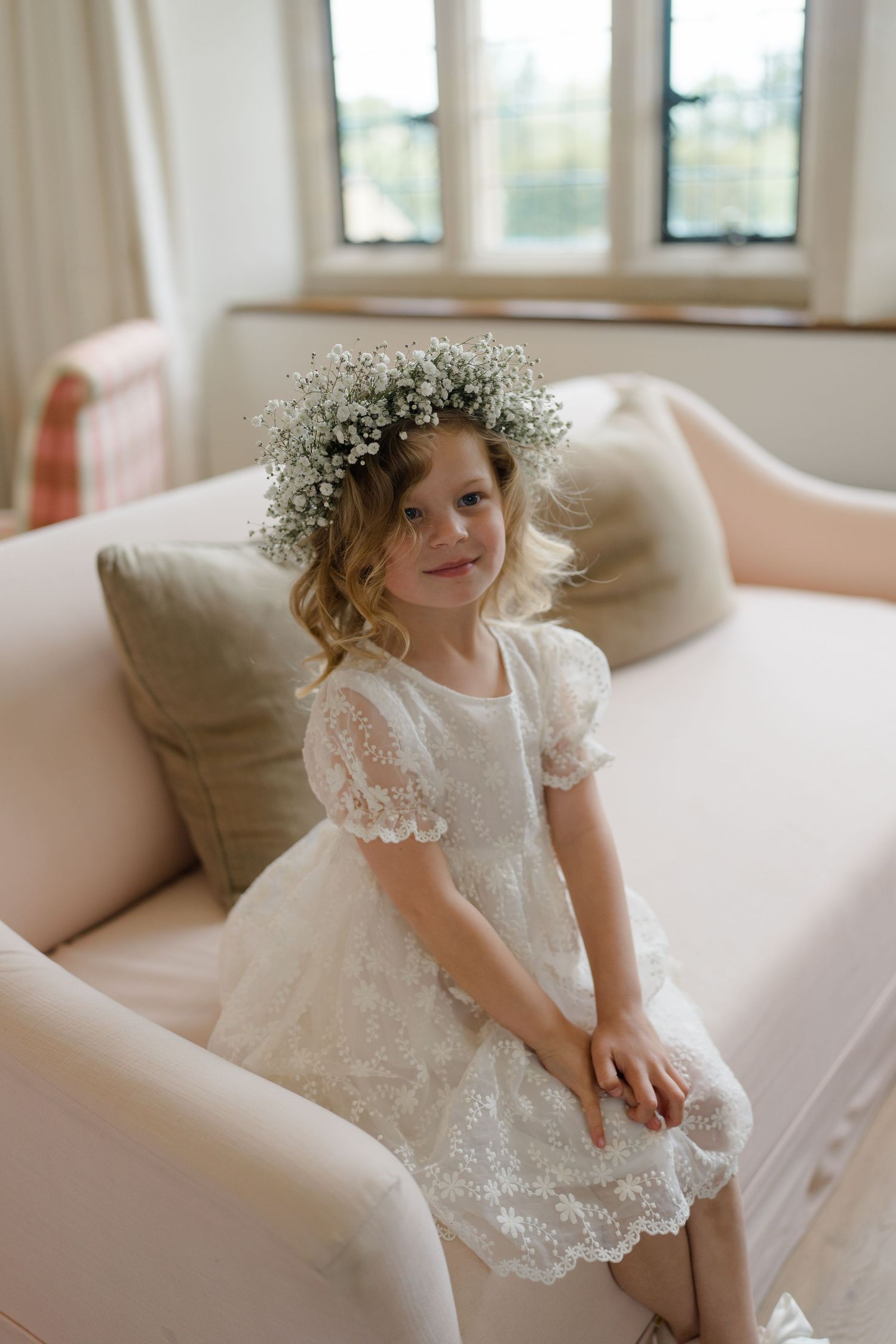 Adorable flower girl in white lace dress wearing baby's breath floral crown seated on sofa.