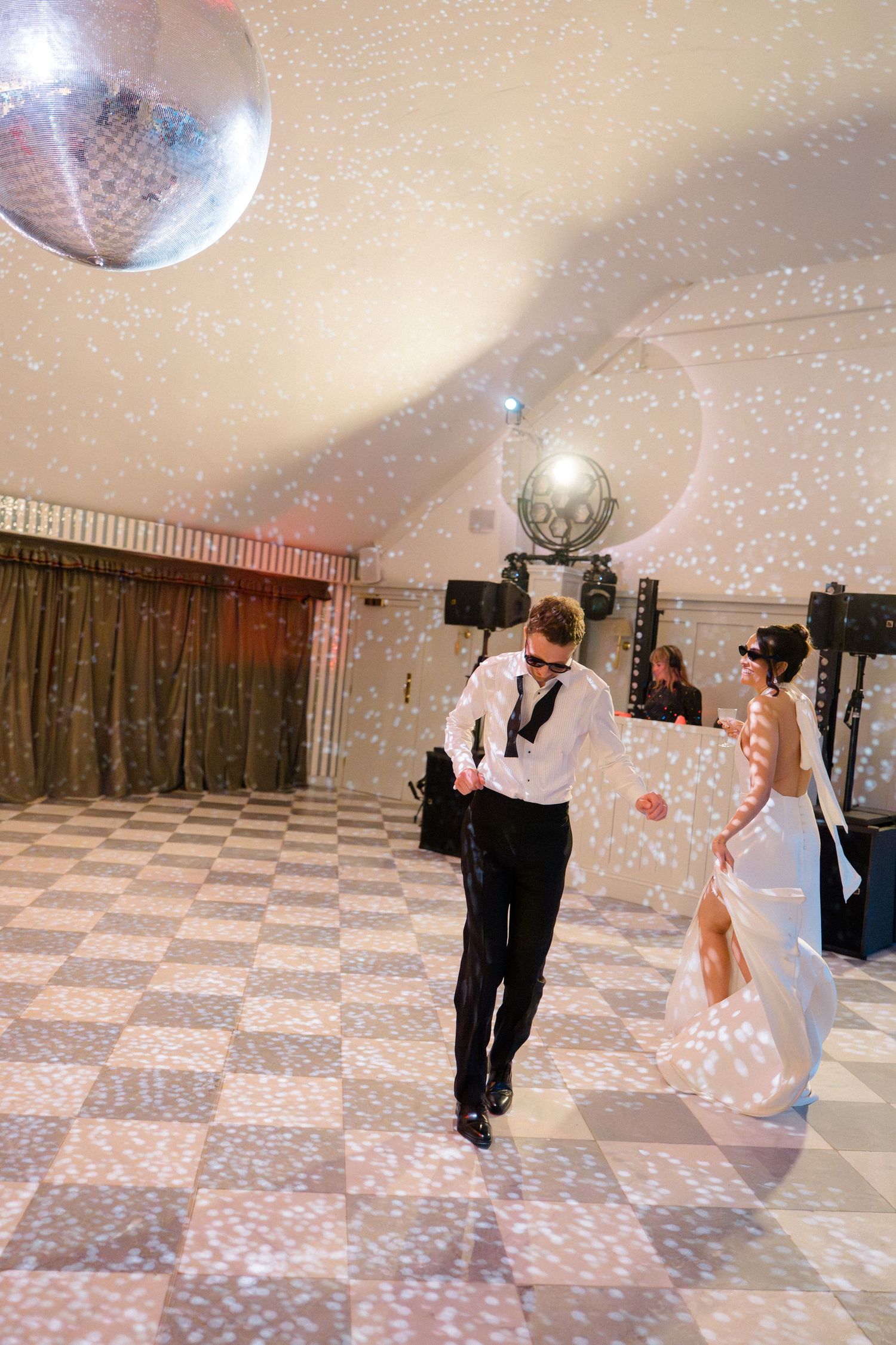 Groom dances energetically with bride in backless gown under glittering disco ball on checkered wedding dance floor.