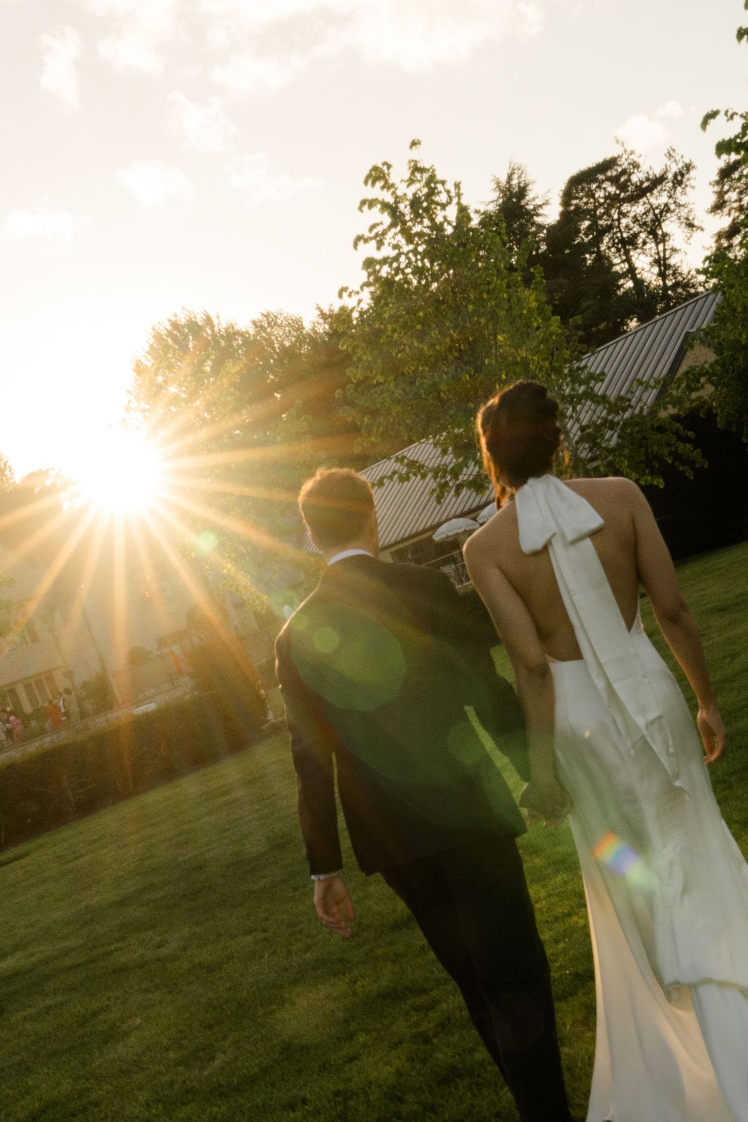 Bride in backless white gown and groom in black suit walk toward golden sunset across lush green lawn.