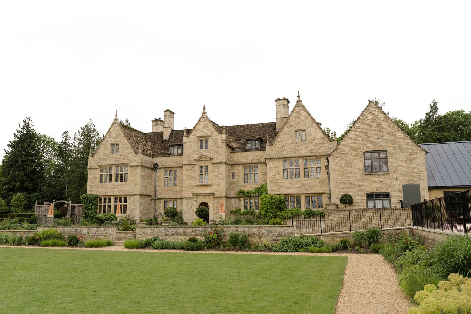 A grand Cotswold stone manor house with manicured gardens and lawns surrounded by mature trees on an overcast day.
