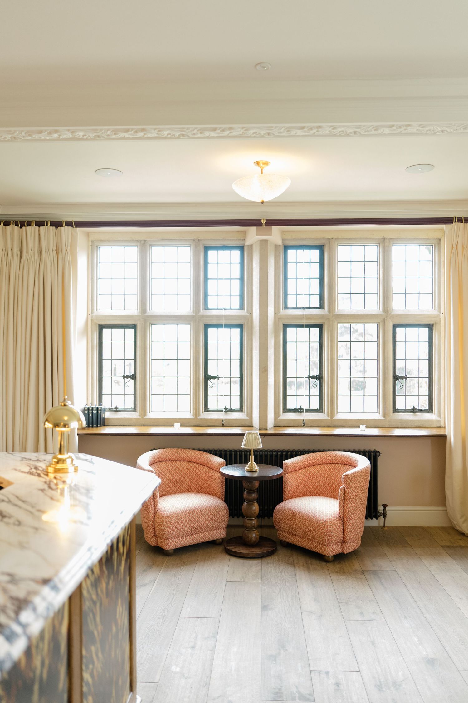 Elegant sitting area with two pink velvet armchairs beside large leaded glass bay windows in a bright hotel room.