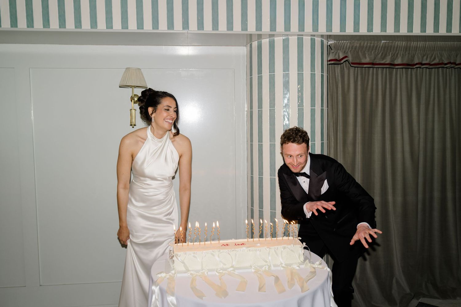 Bride laughs as groom dramatically leans over tiered wedding cake covered in candles at reception.