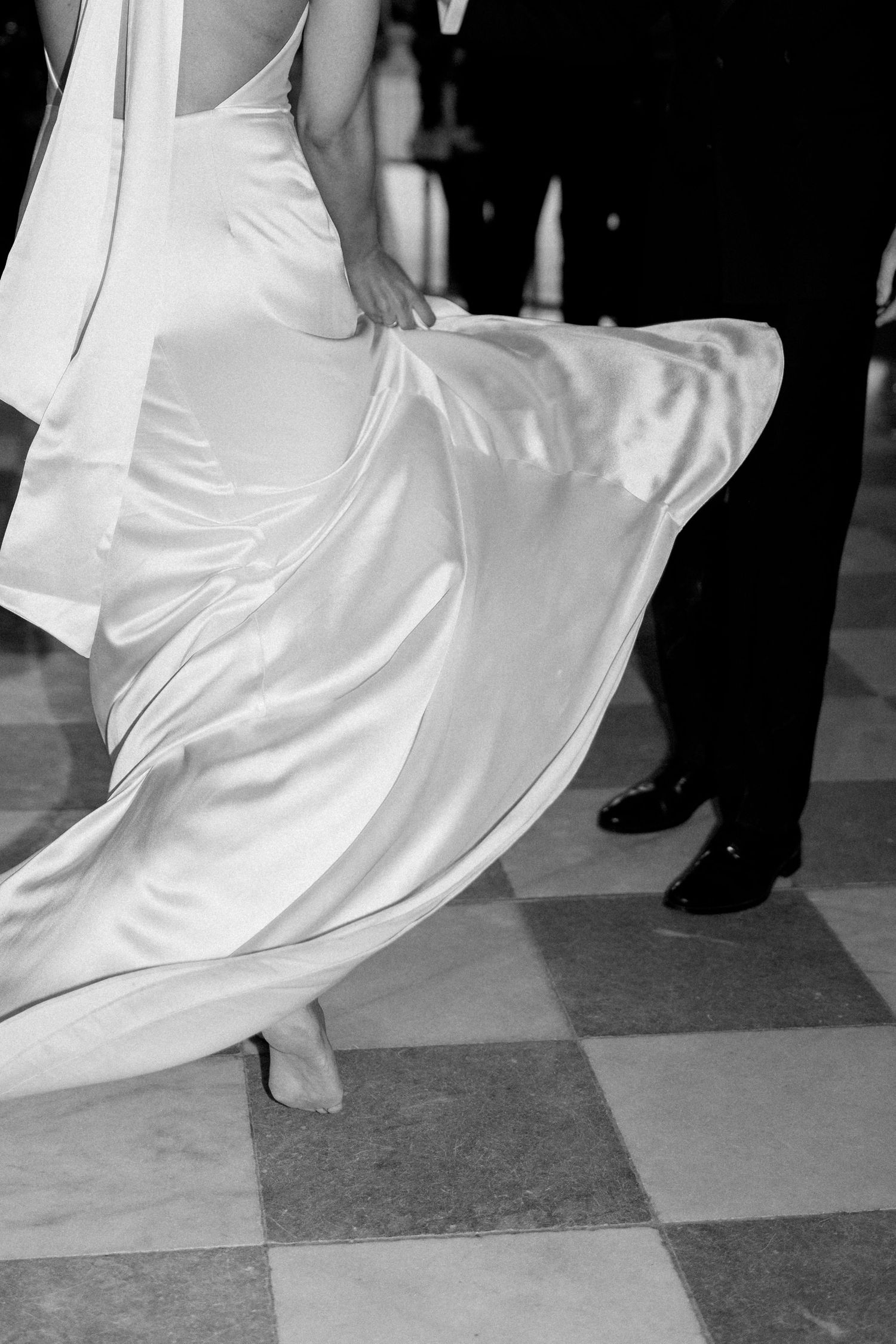 Black and white close-up of a bride twirling her satin wedding dress on a checkered dance floor.
