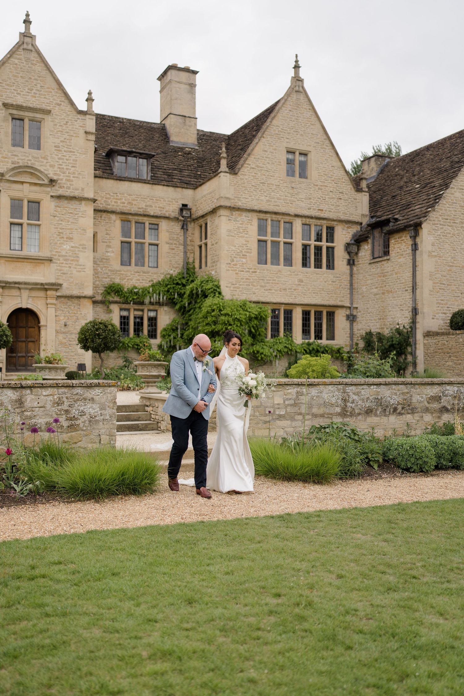 Bride in white gown walks with escort through gardens of a stunning Cotswolds stone manor house.