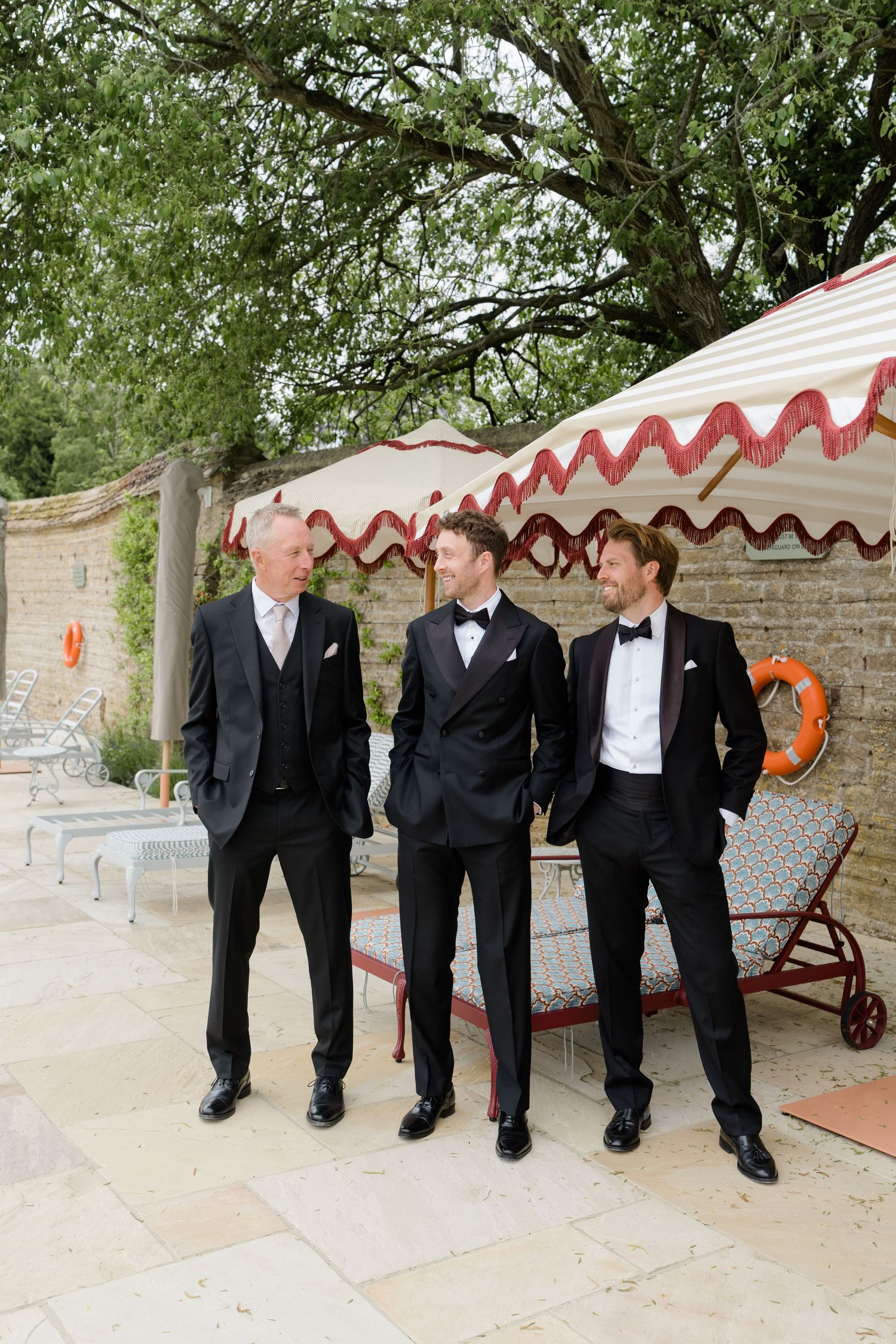 Three men in black tuxedos with bow ties conversing beside a striped poolside cabana.