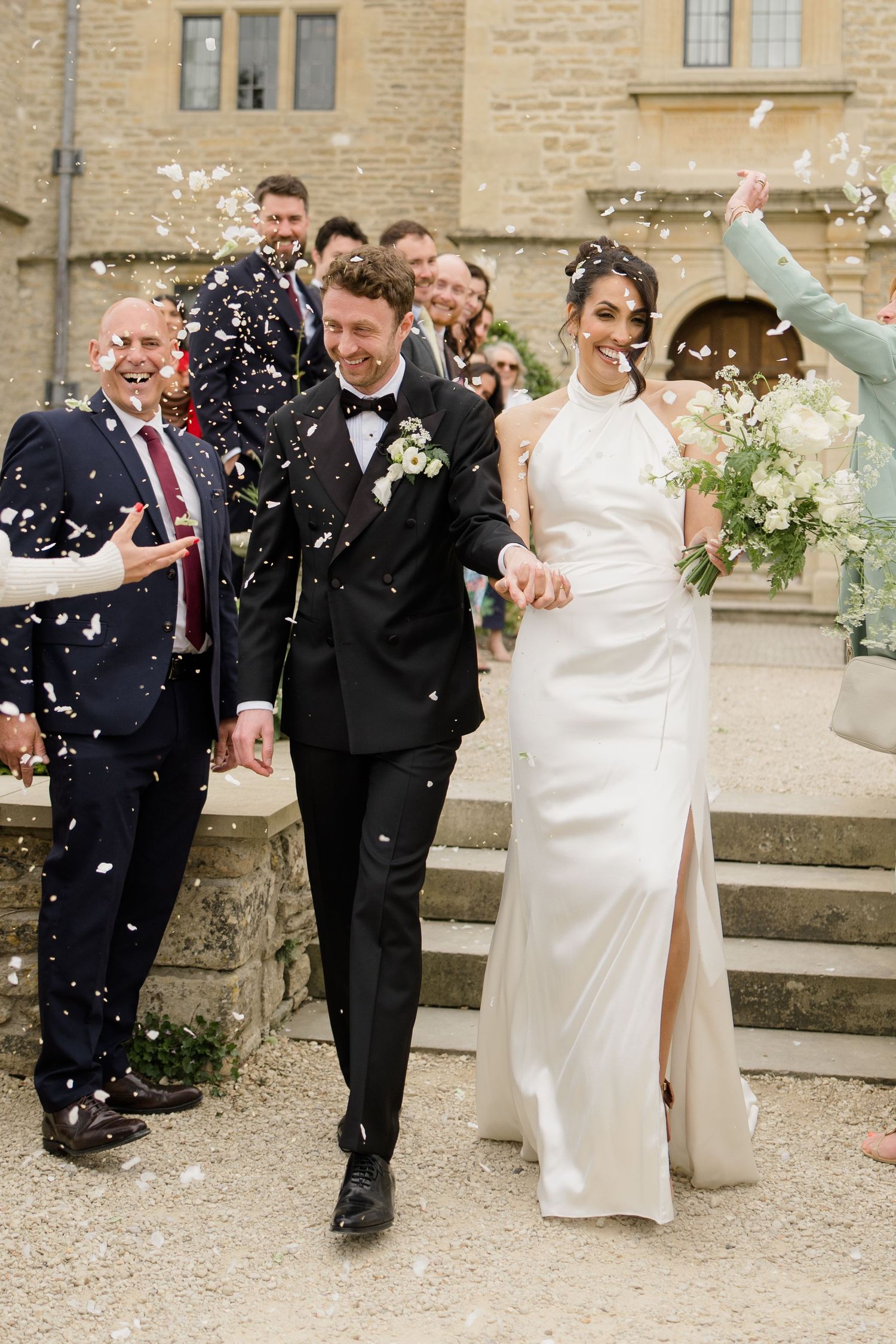 Joyful newlyweds walk through confetti shower outside stone manor house surrounded by cheering wedding guests.