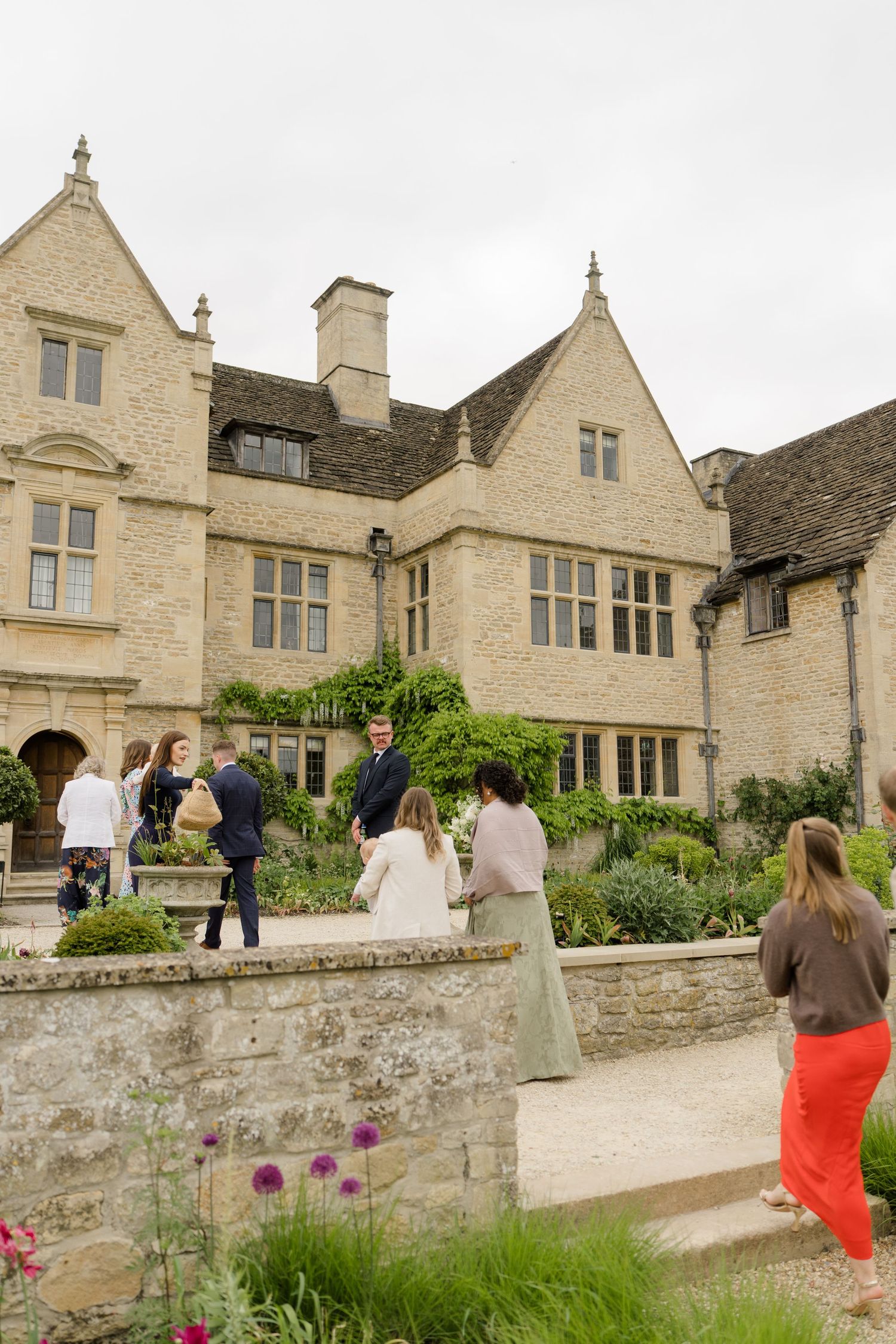 Wedding guests mingling outside a grand Cotswolds stone manor house surrounded by lush garden flowers.