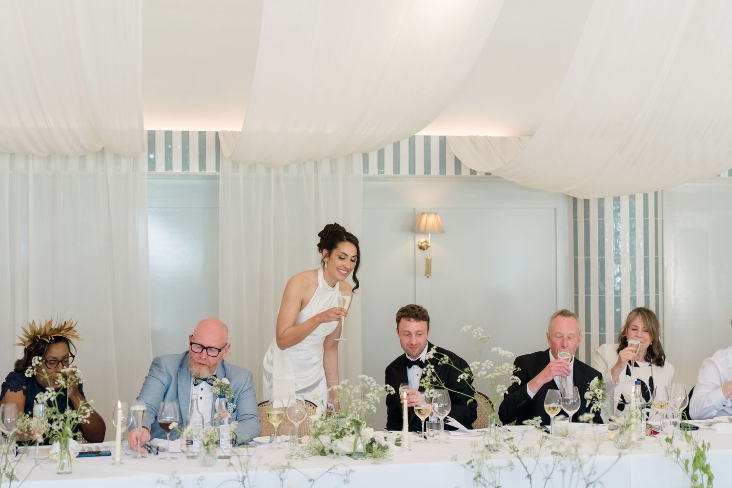 Bride delivers speech at wedding top table surrounded by smiling guests in elegant marquee.