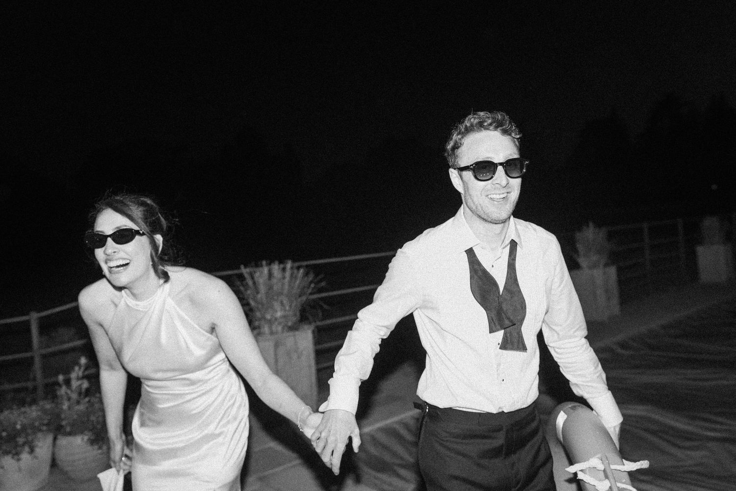 Joyful bride and groom holding hands wearing sunglasses in black and white night photo.