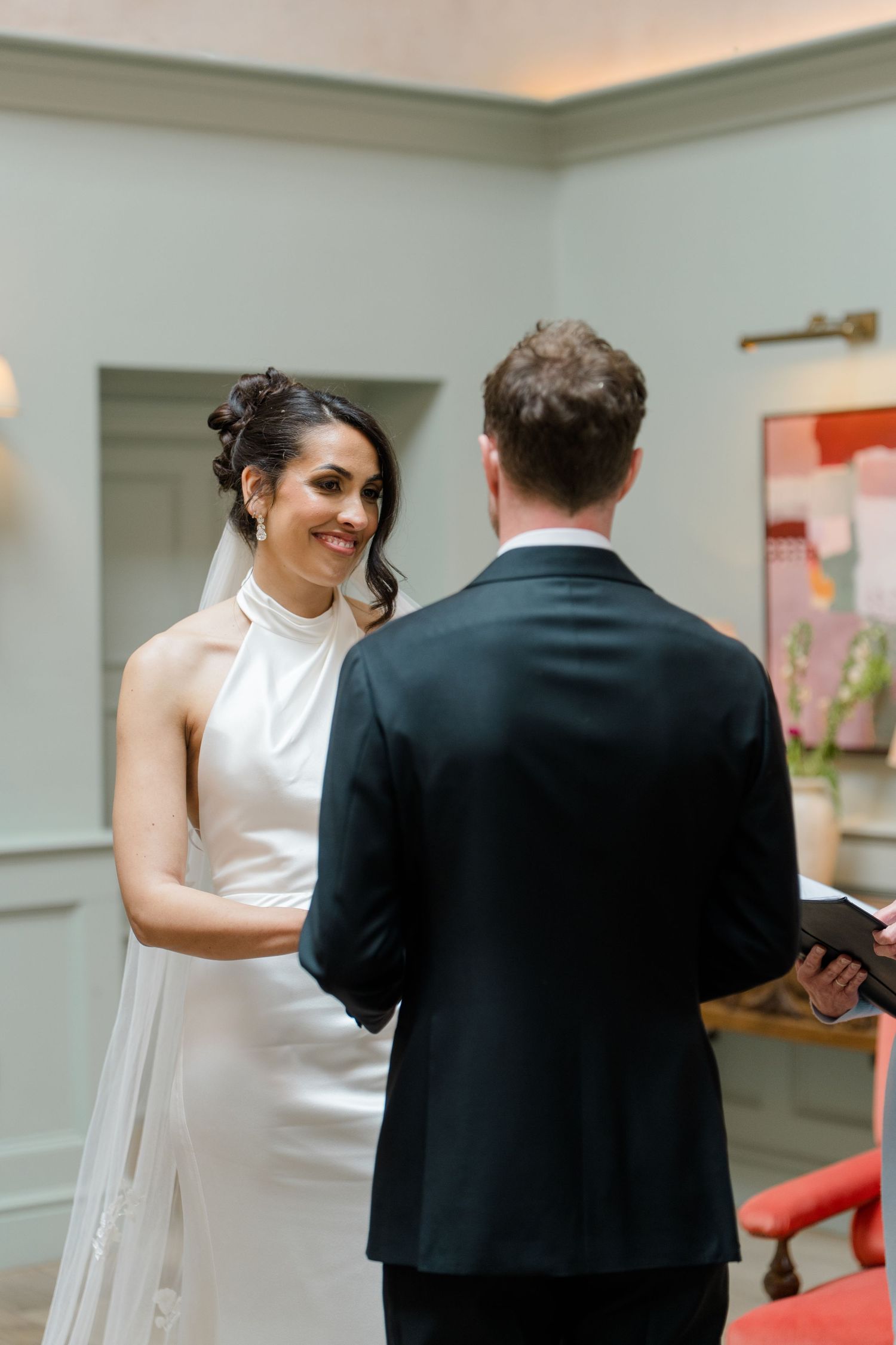 Bride in halter neck gown smiles at groom during intimate indoor wedding ceremony vow exchange.