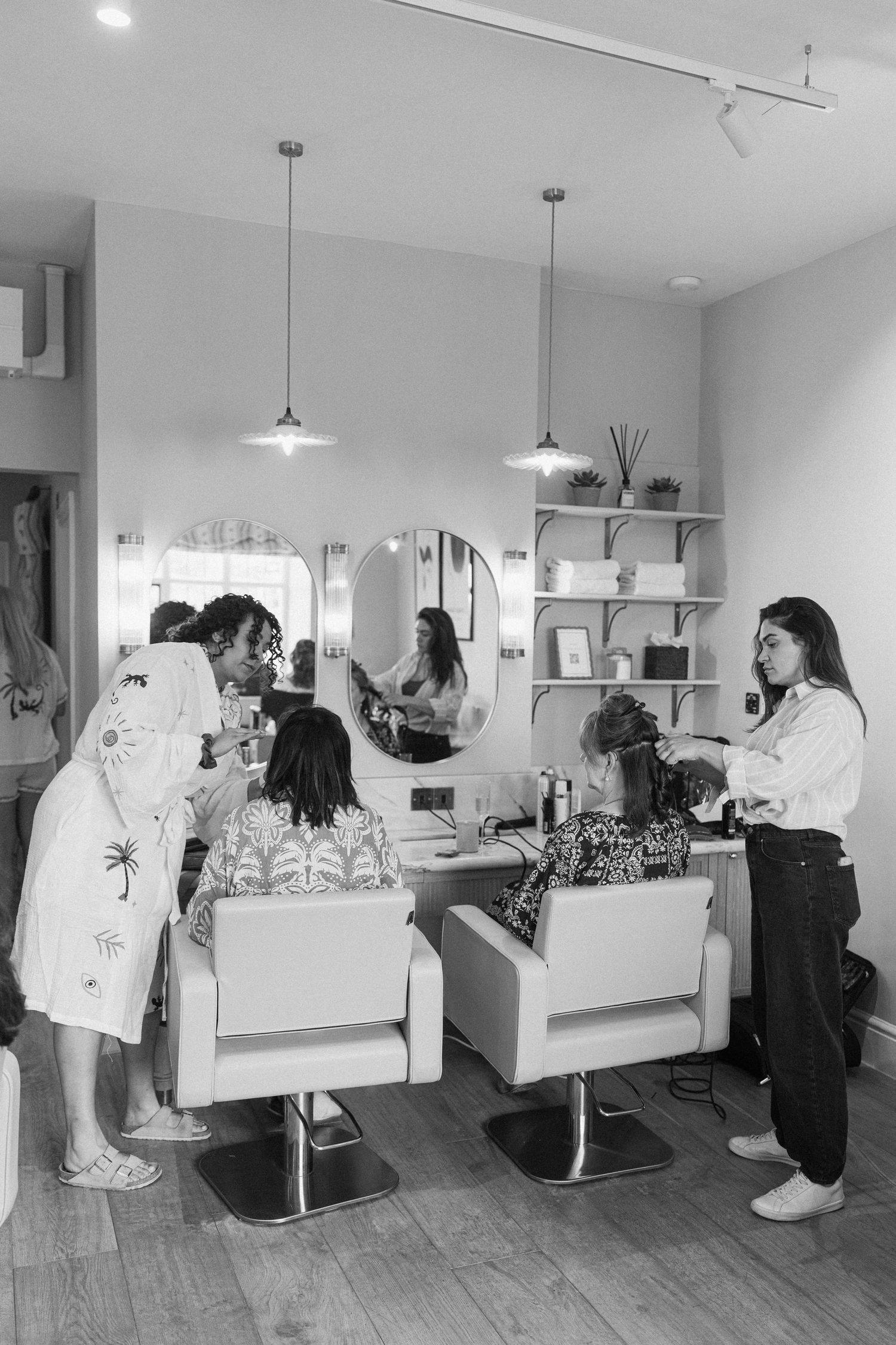Black and white photo of bridal party getting hair styled in a modern salon with pendant lights.
