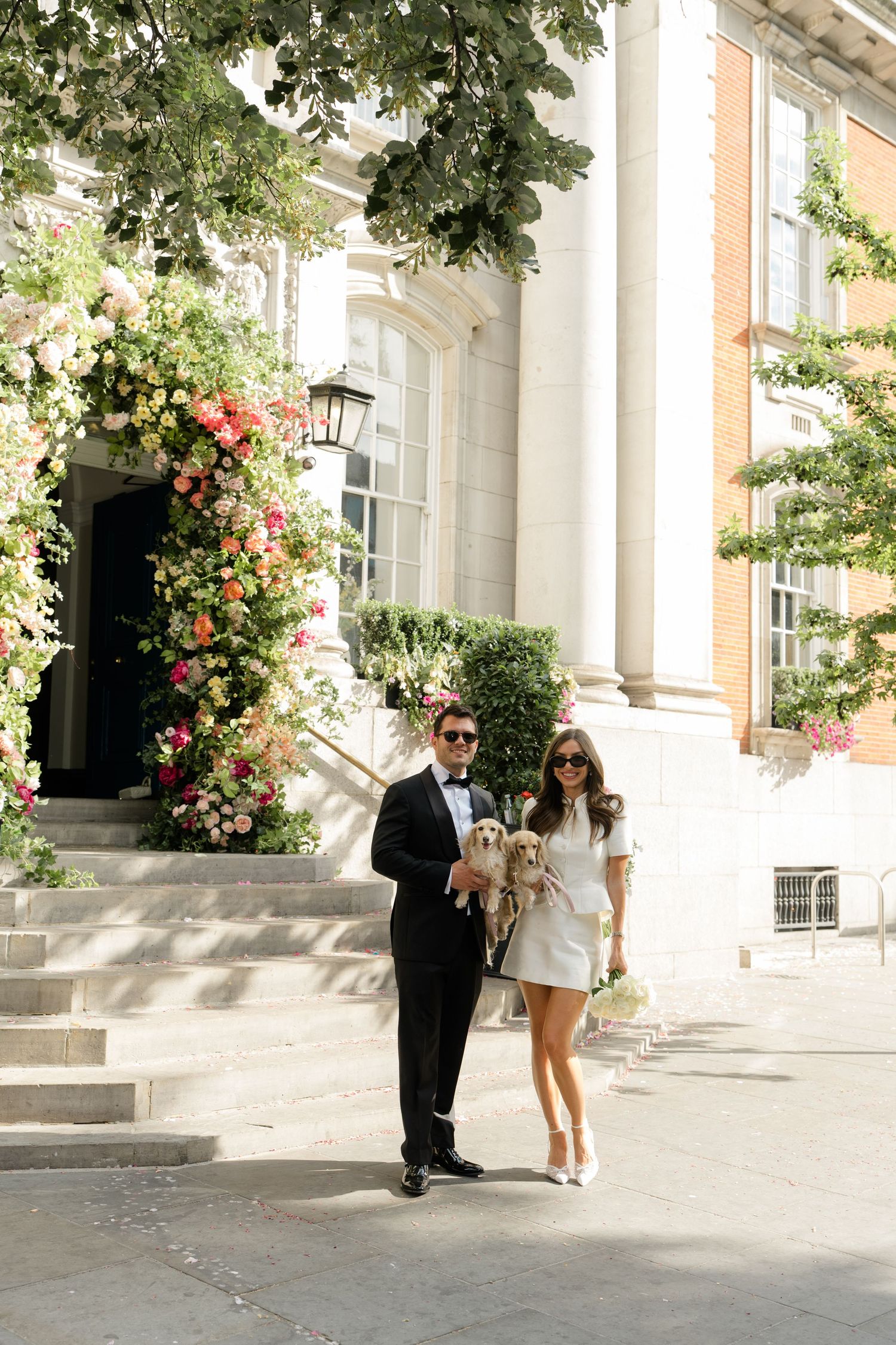Smiling couple in wedding attire holding a small dog outside a floral-adorned London venue.