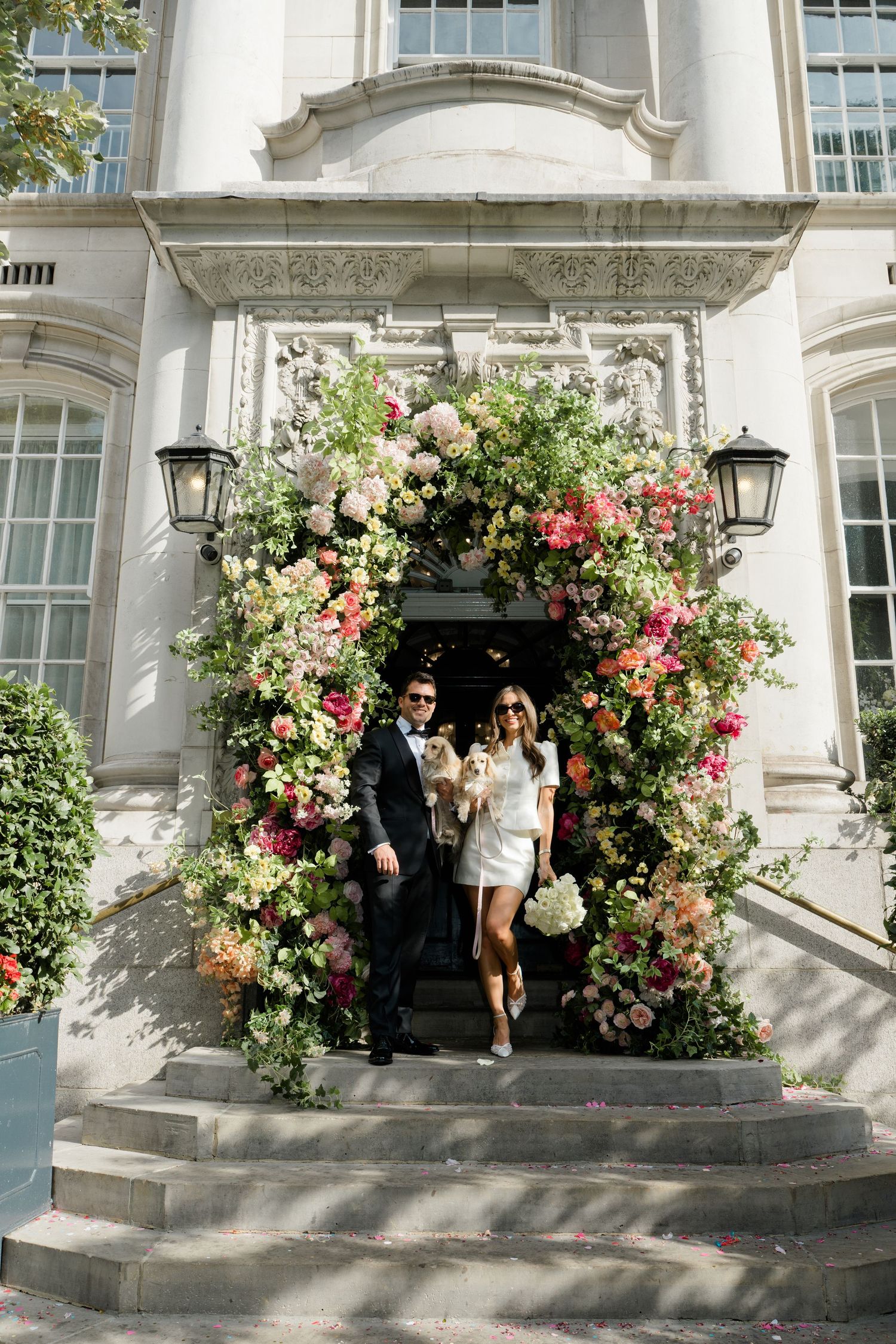 Couple in wedding attire poses under a stunning floral arch at an elegant white stone building entrance.