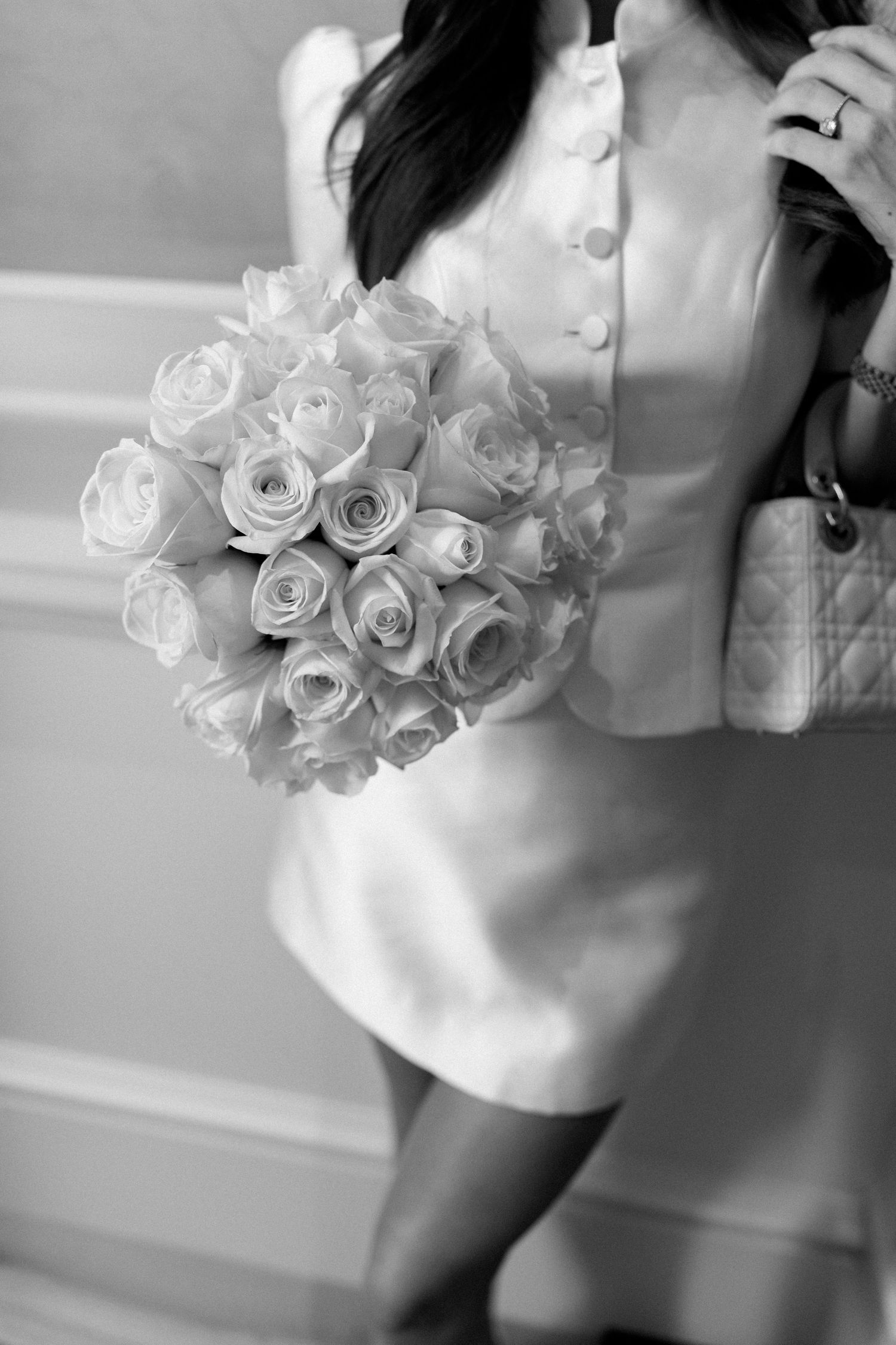 Black and white photo of bride in white mini dress holding a bouquet of roses with a designer handbag.
