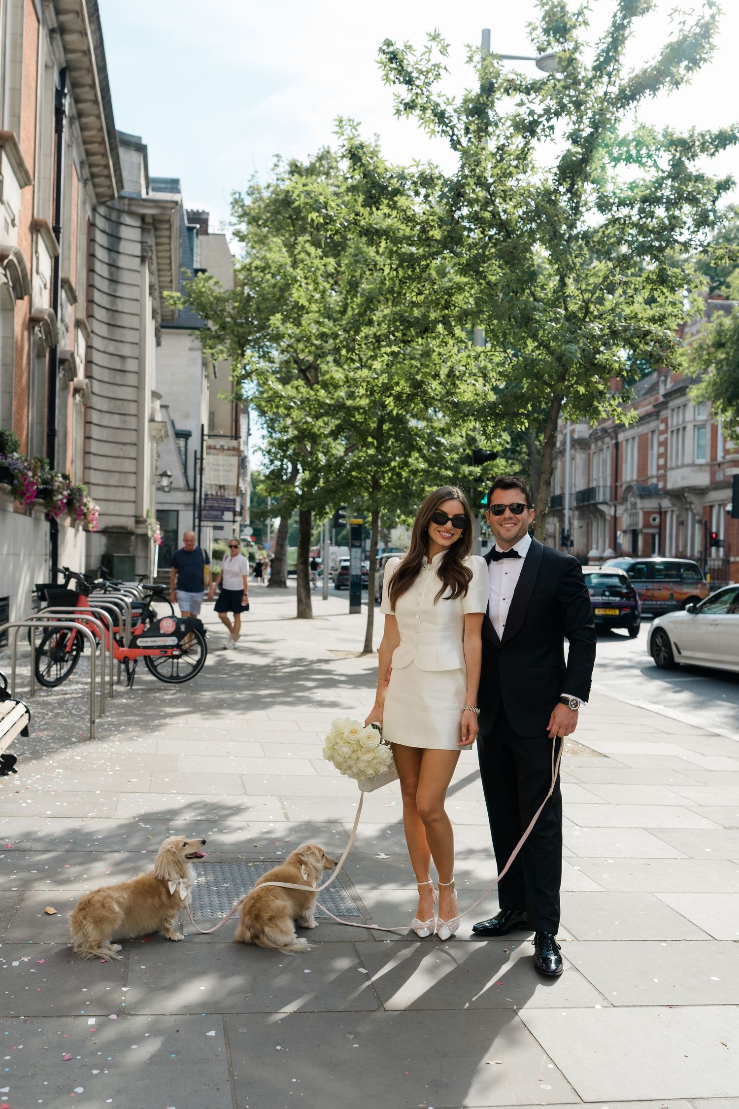 Smiling bride and groom in sunglasses walk two golden dogs on a sunny London street.