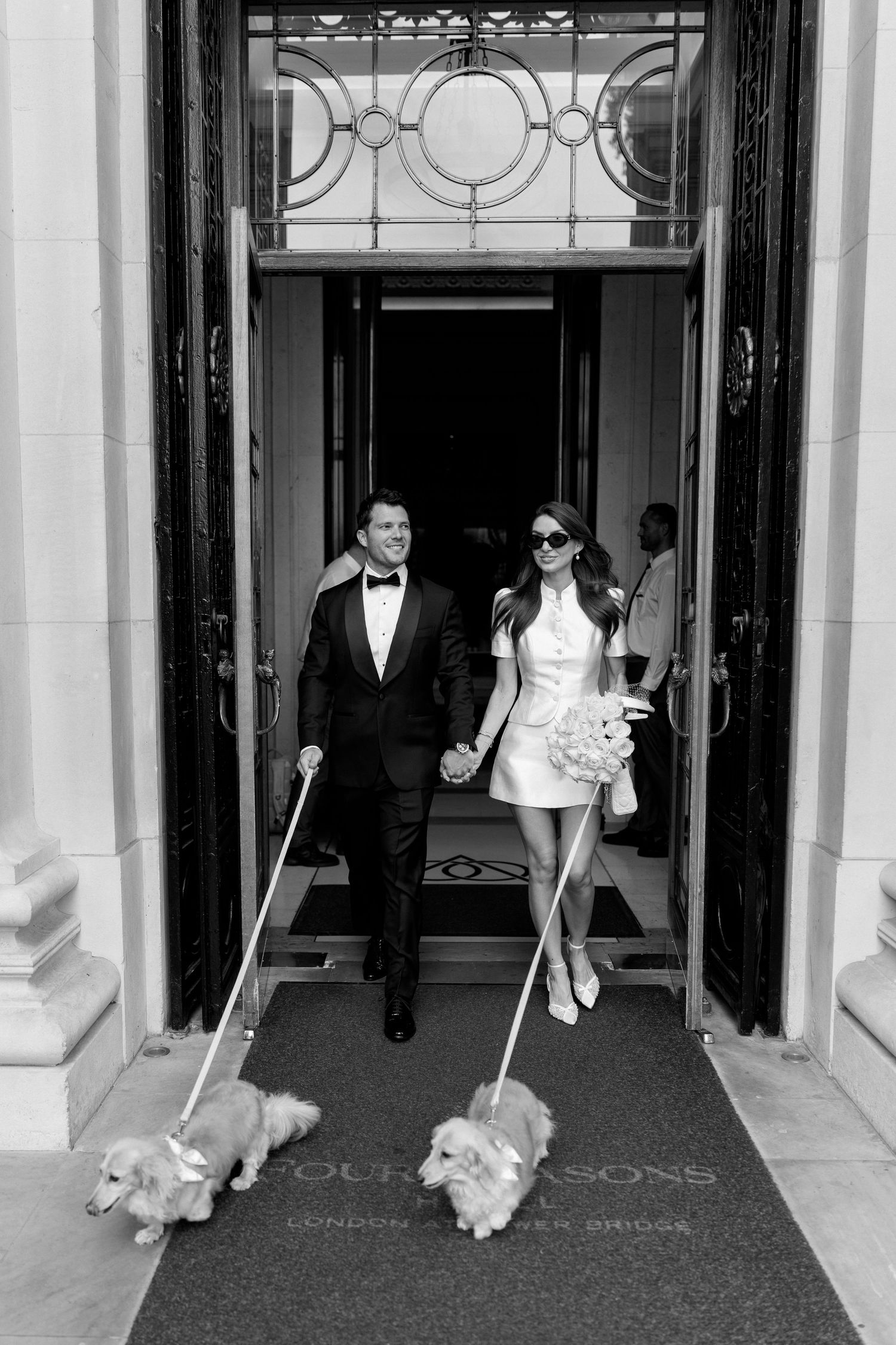 Bride and groom exit Four Seasons London Tower Bridge in black and white, holding hands and walking two Corgis.