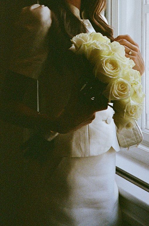 Bride holding a bouquet of white roses near a window, wearing a white wedding dress and jacket.