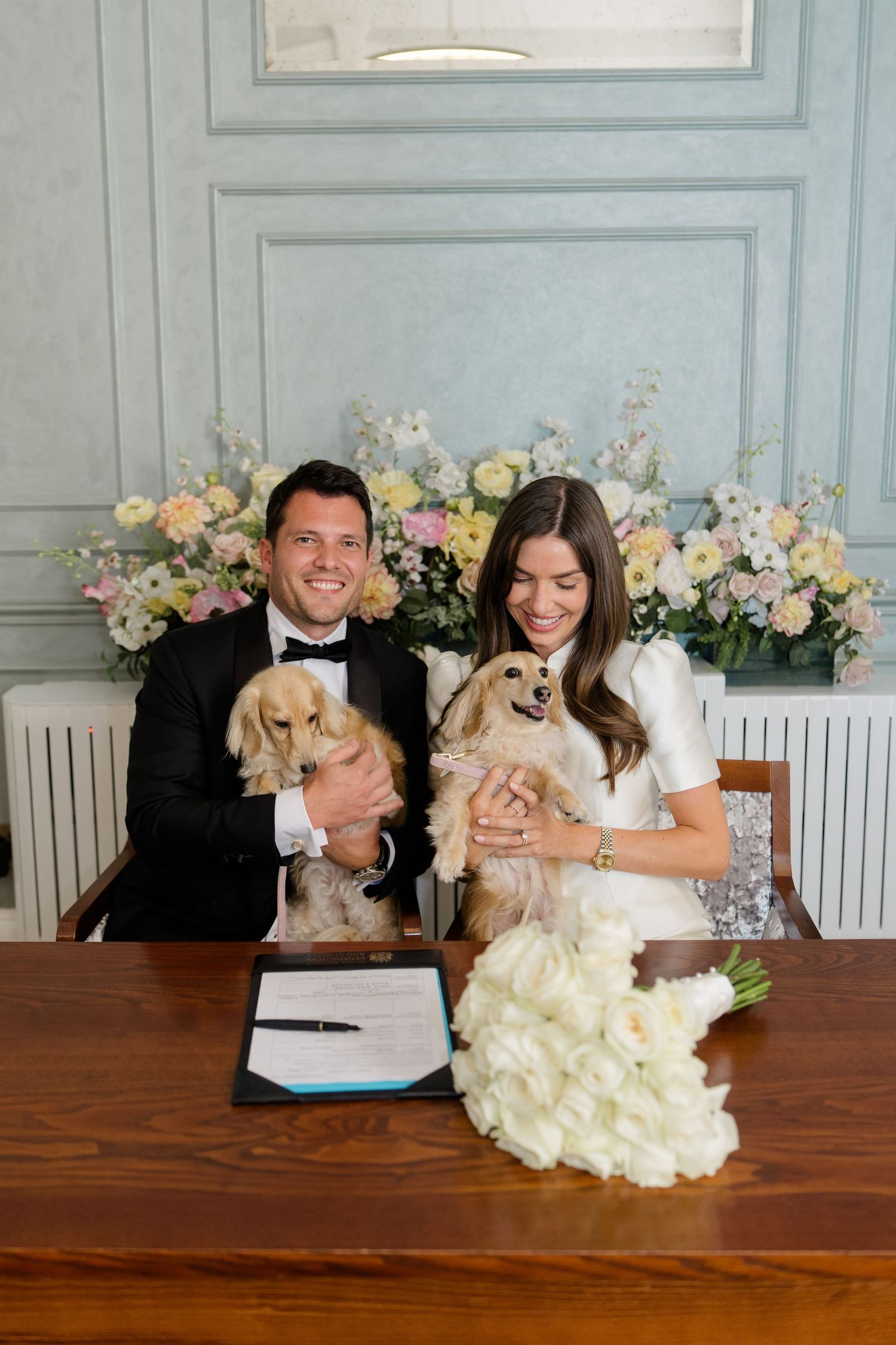 Couple in wedding attire holding two golden Cocker Spaniel puppies at a signing table with white tulip bouquet.