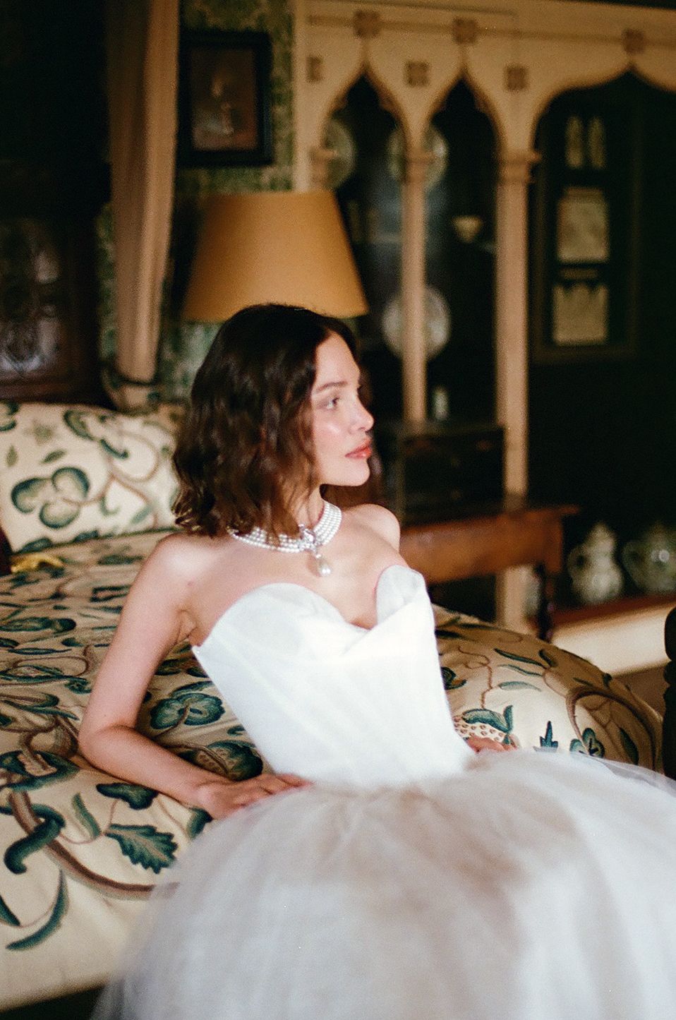 A person in a white wedding dress reclines on a patterned couch in an elegant interior setting.