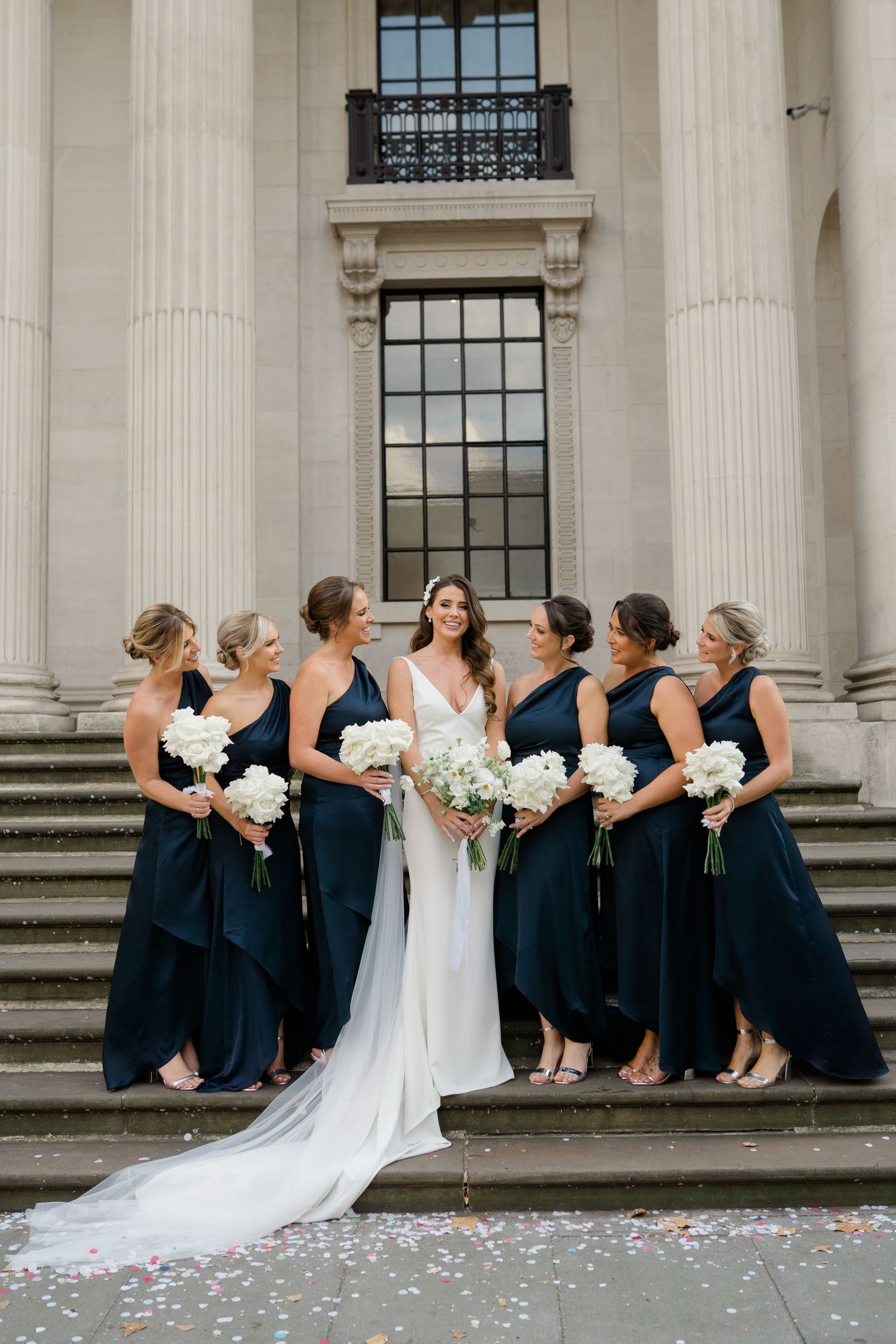 Bridesmaids in navy dresses line up with bride in white gown against classical stone building.