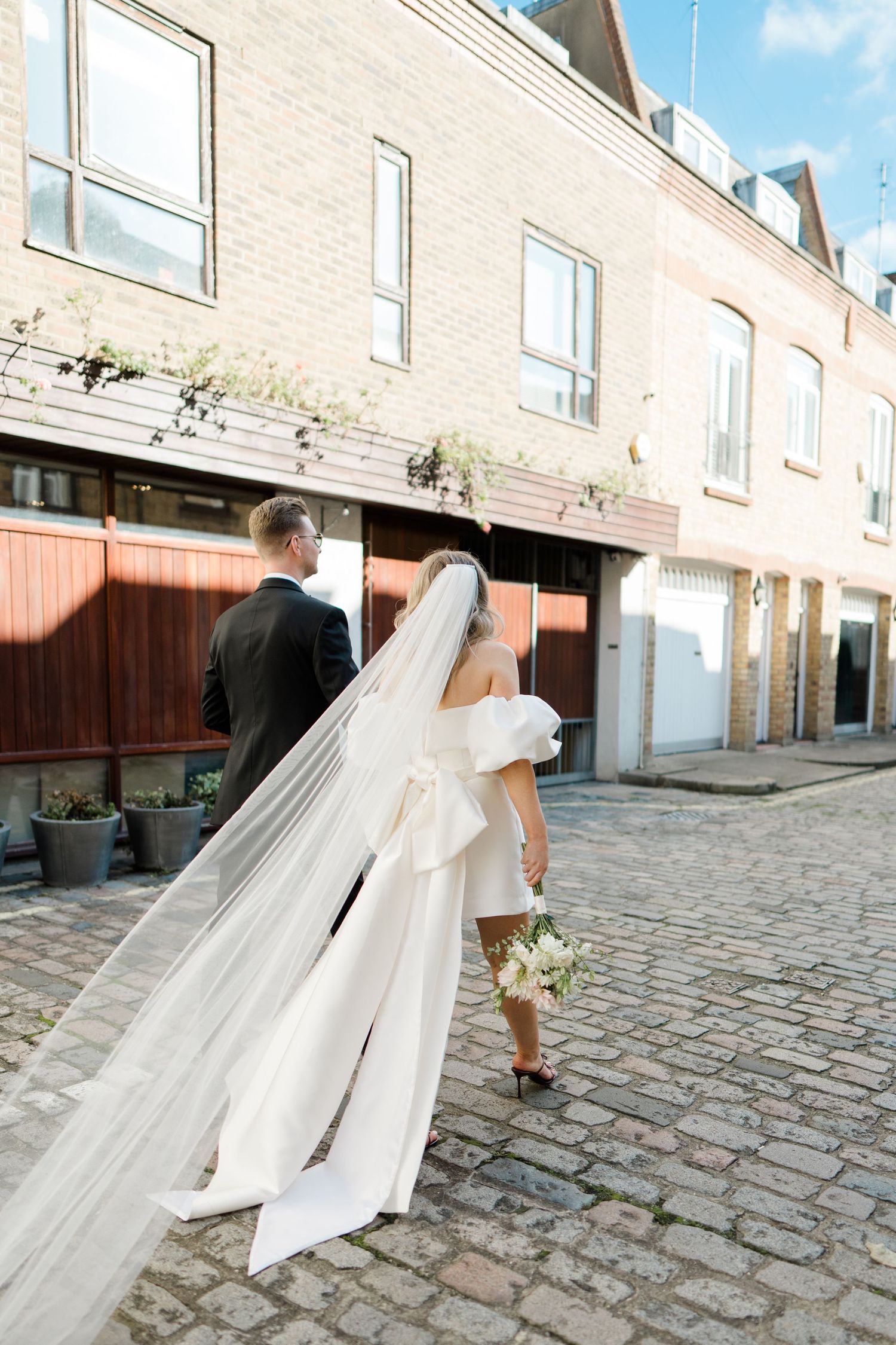A wedding couple walks down a cobblestone street with the bride's long veil trailing behind them.