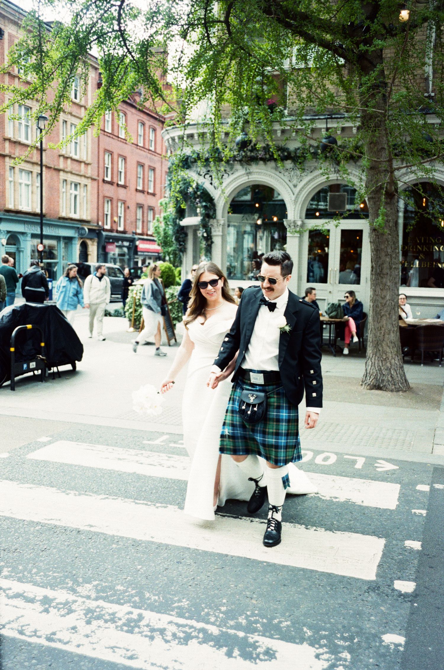 A couple walking across a crosswalk in Edinburgh, with the man wearing a traditional Scottish kilt and formal jacket.