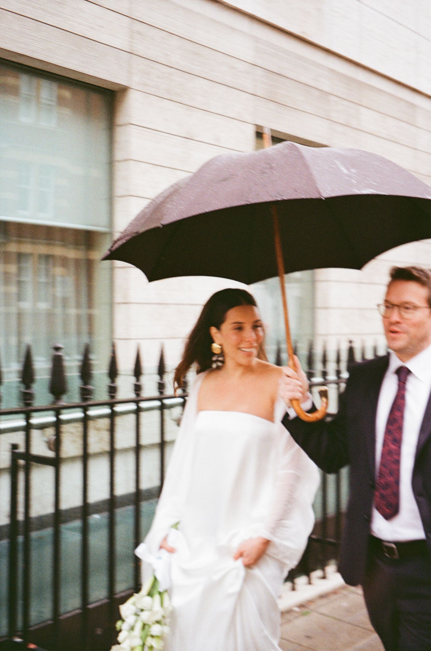 A wedding couple shares an umbrella while walking outside a building with white stone walls on a rainy day.