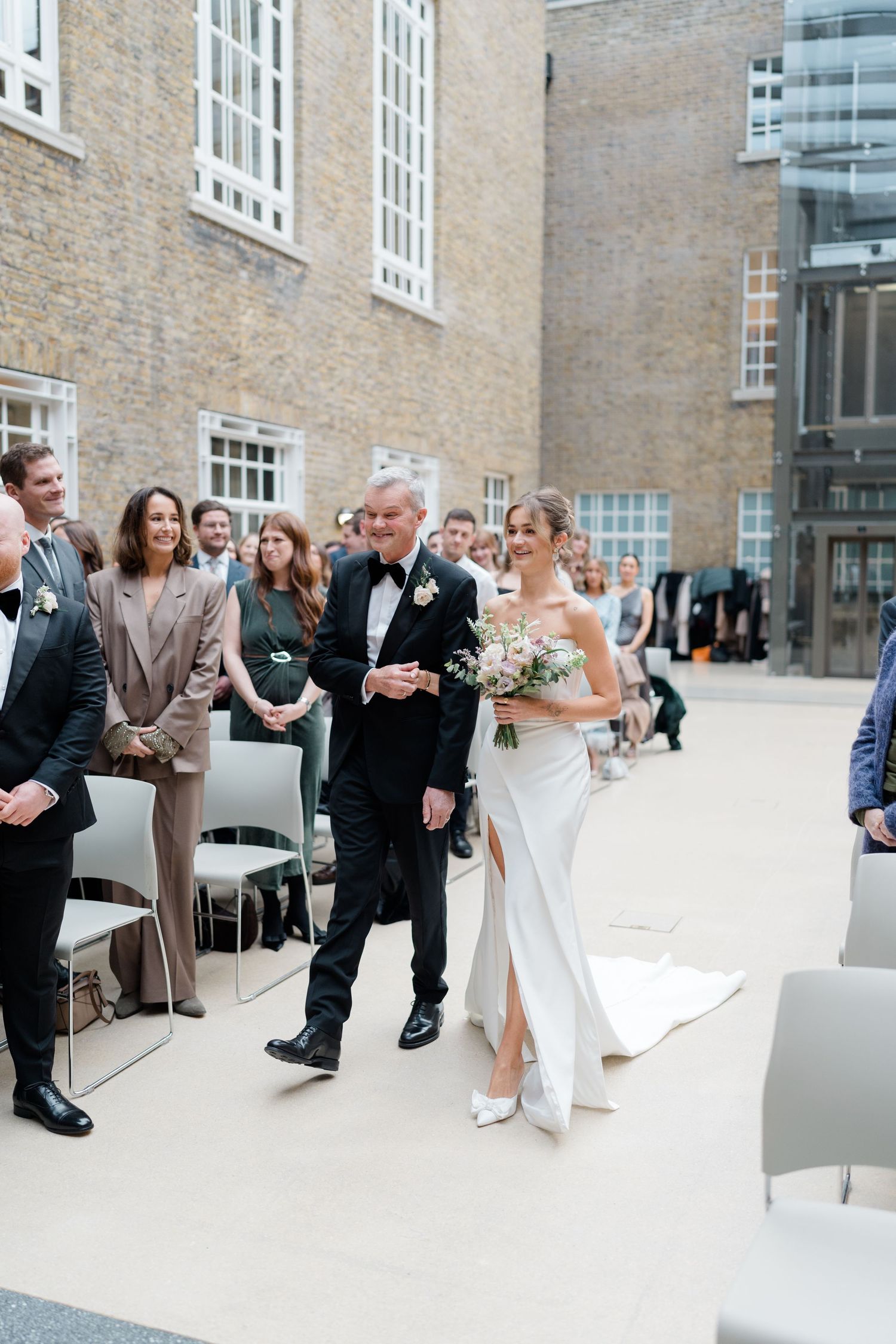 Elegant outdoor wedding ceremony with guests seated in white chairs against a stone building facade.