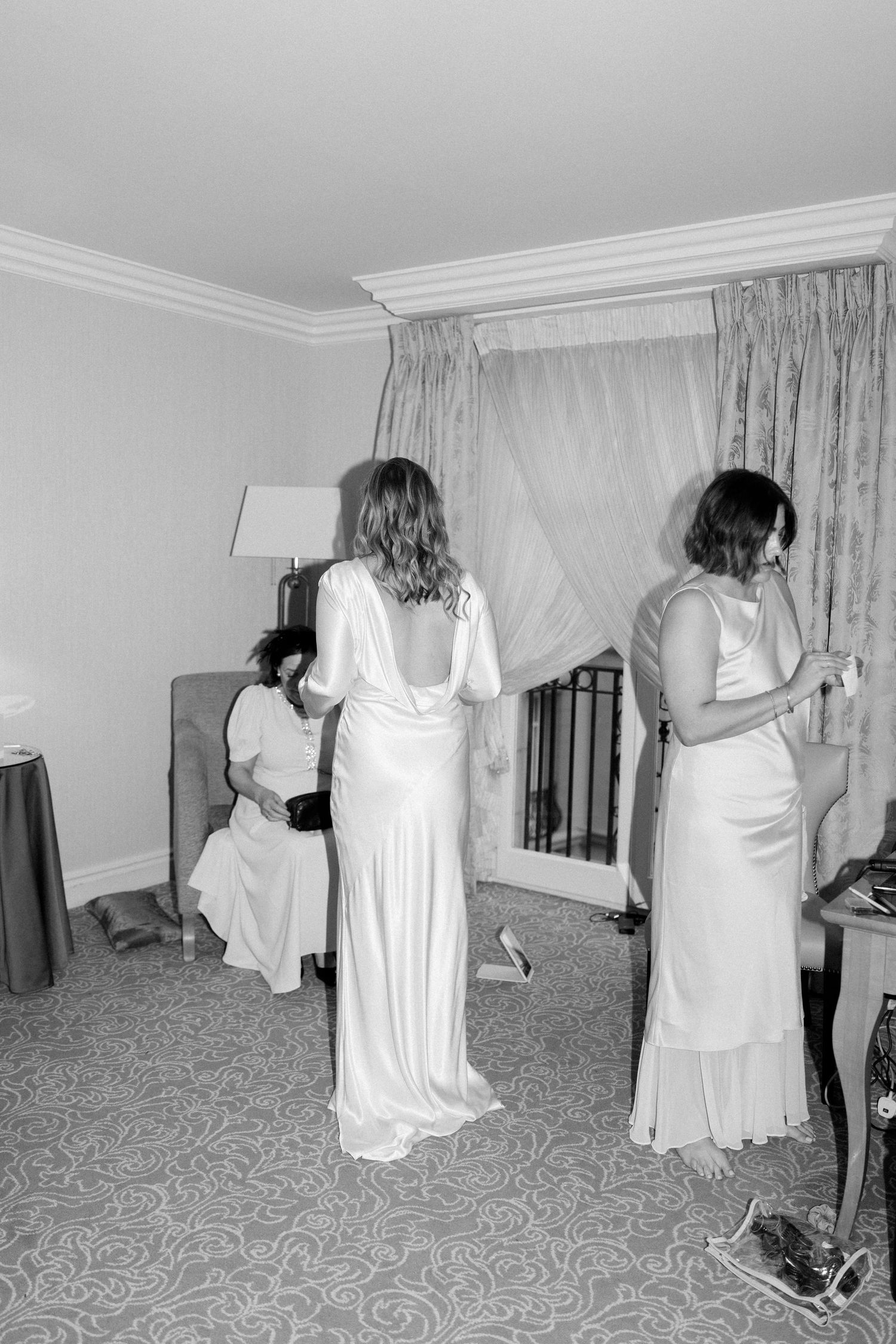 Black and white photo series showing white dresses being fitted in an elegant hotel room with patterned carpet.