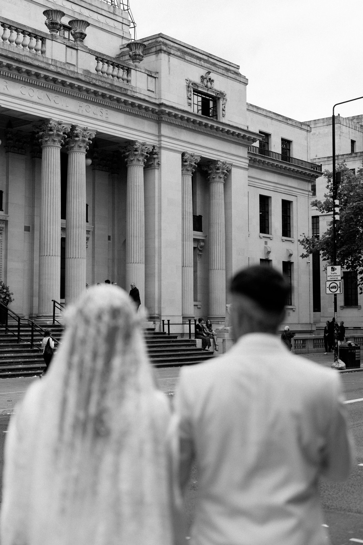 A couple views a classical building with columns from behind in black and white.