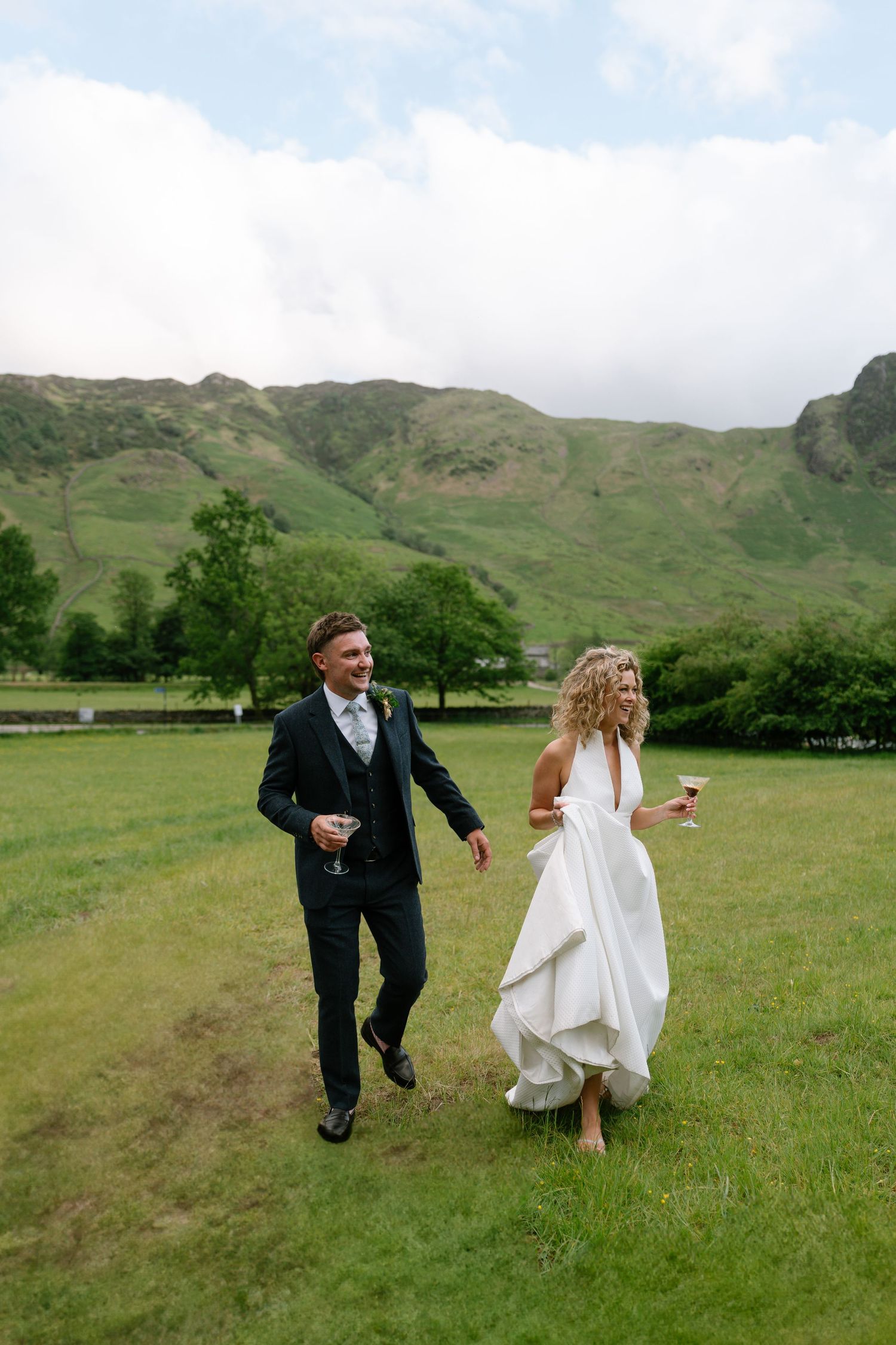 A couple playfully runs through a green field with mountains in the backdrop.