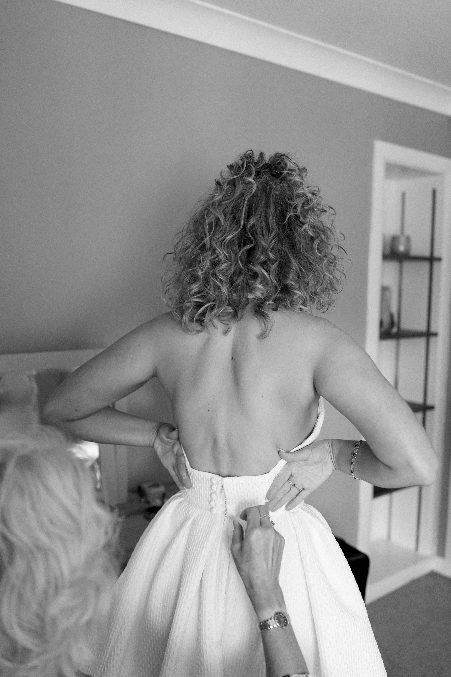 Black and white photo of someone getting their wedding dress buttoned up, showing curly hair and bare shoulders.