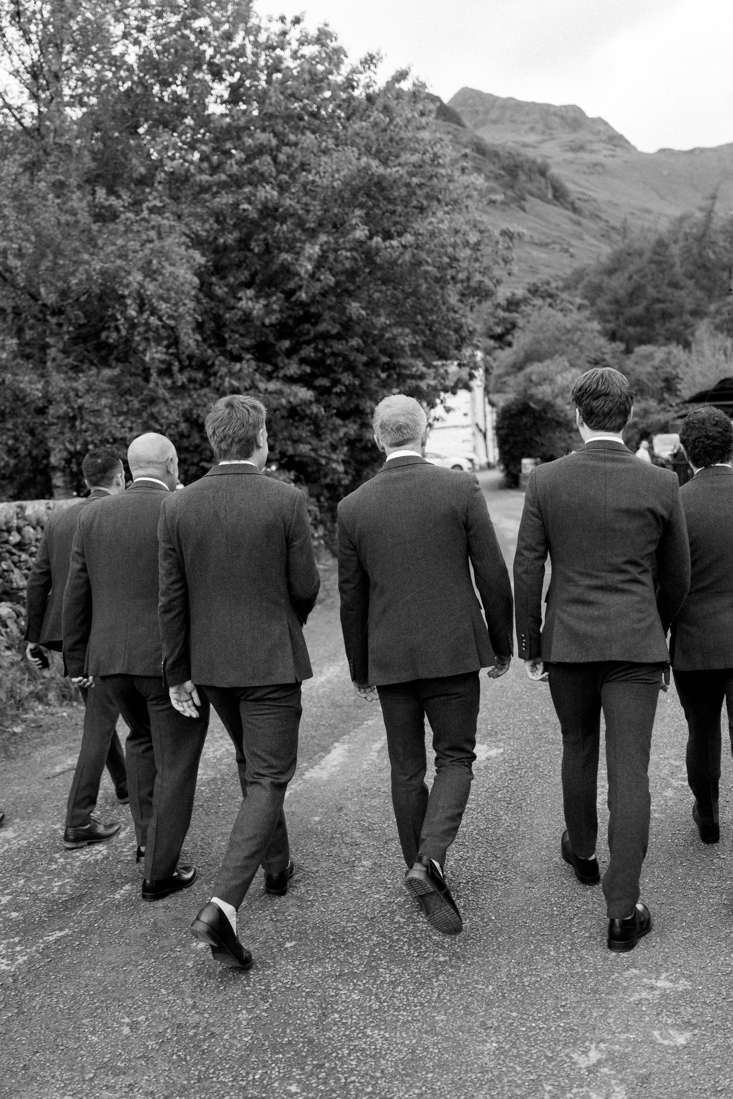 A group of groomsmen in dark suits walking together on a path with mountains in the background.