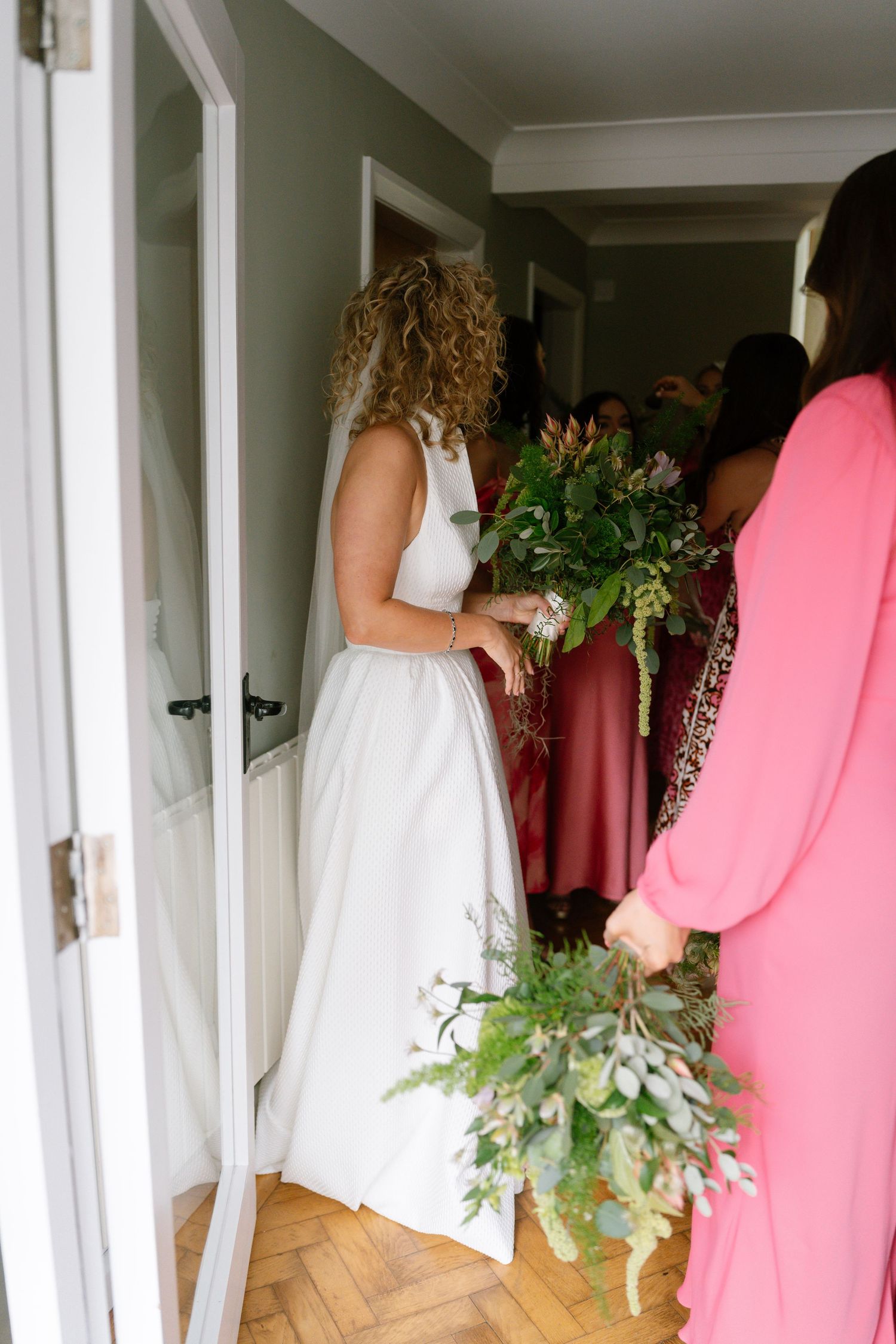 Wedding party in pink dresses and white gown walking through interior hallway.