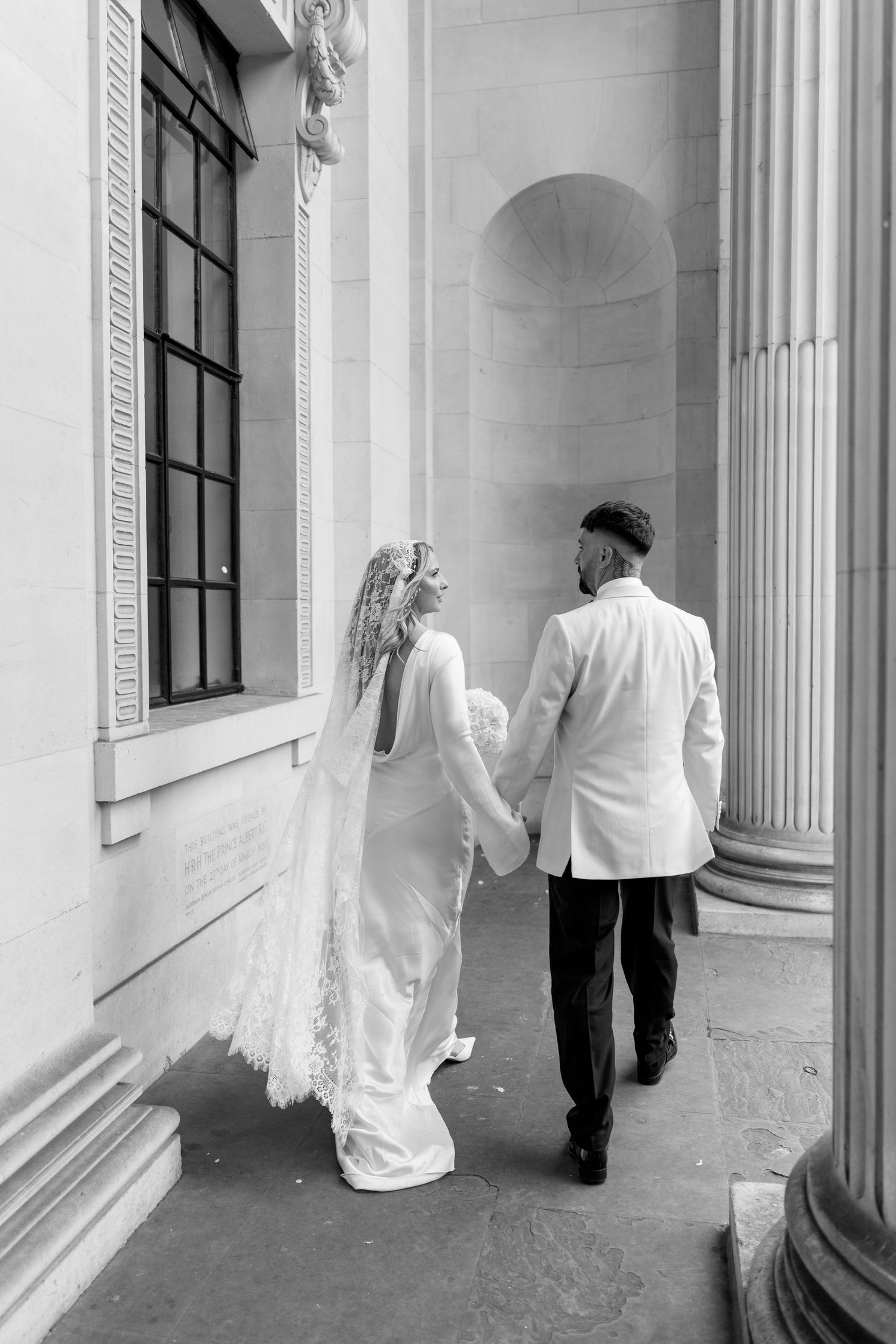 A black and white photo of newlyweds walking hand in hand through a classical columned building.