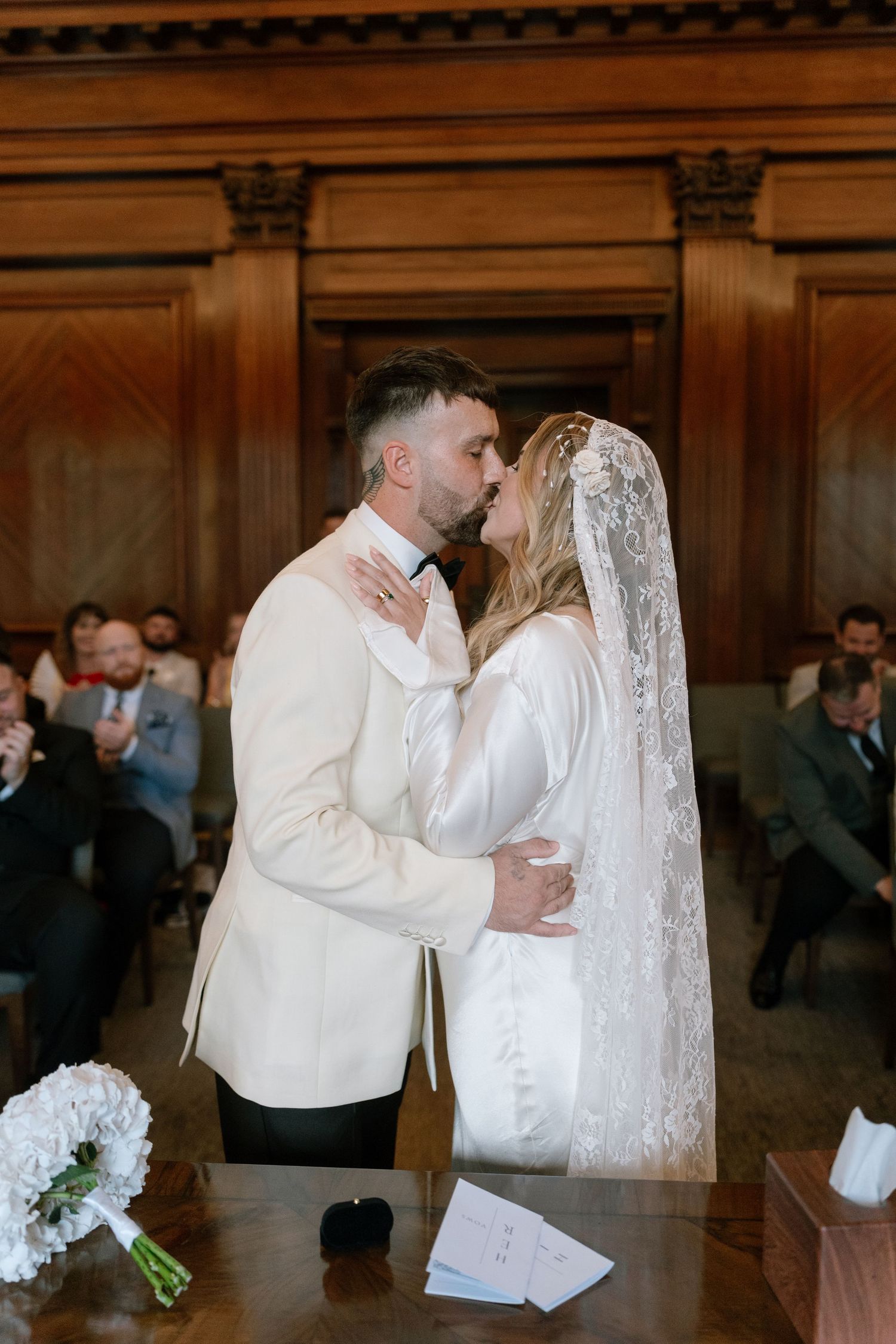 Romantic wedding ceremony moment in a wood-paneled room with guests watching in background.