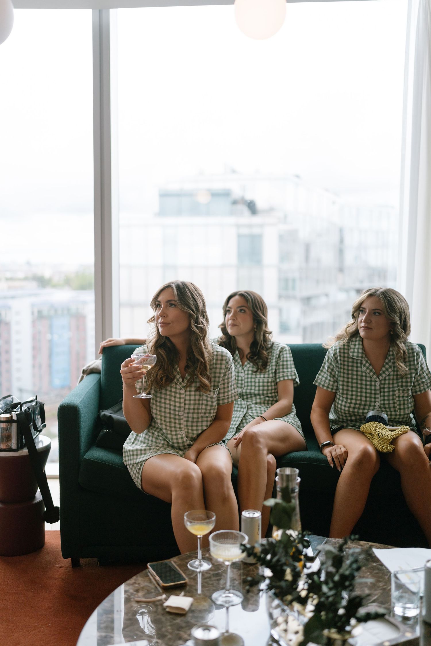 Bridesmaids in matching gingham dresses relaxing on a couch with drinks and city views.
