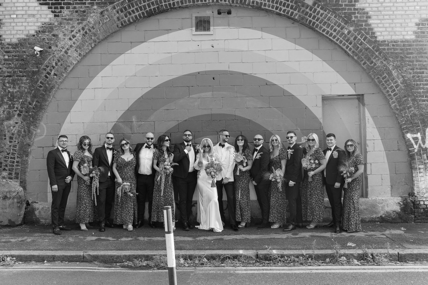 Black and white photo of wedding party posing under brick archway.