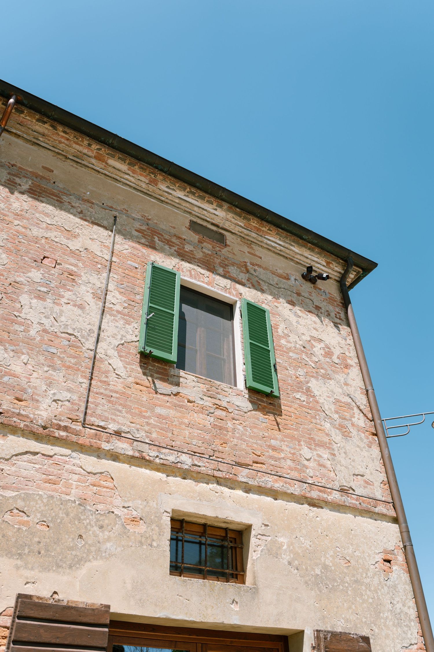 Old Italian stone building with weathered pink walls and green shuttered windows against bright blue sky.